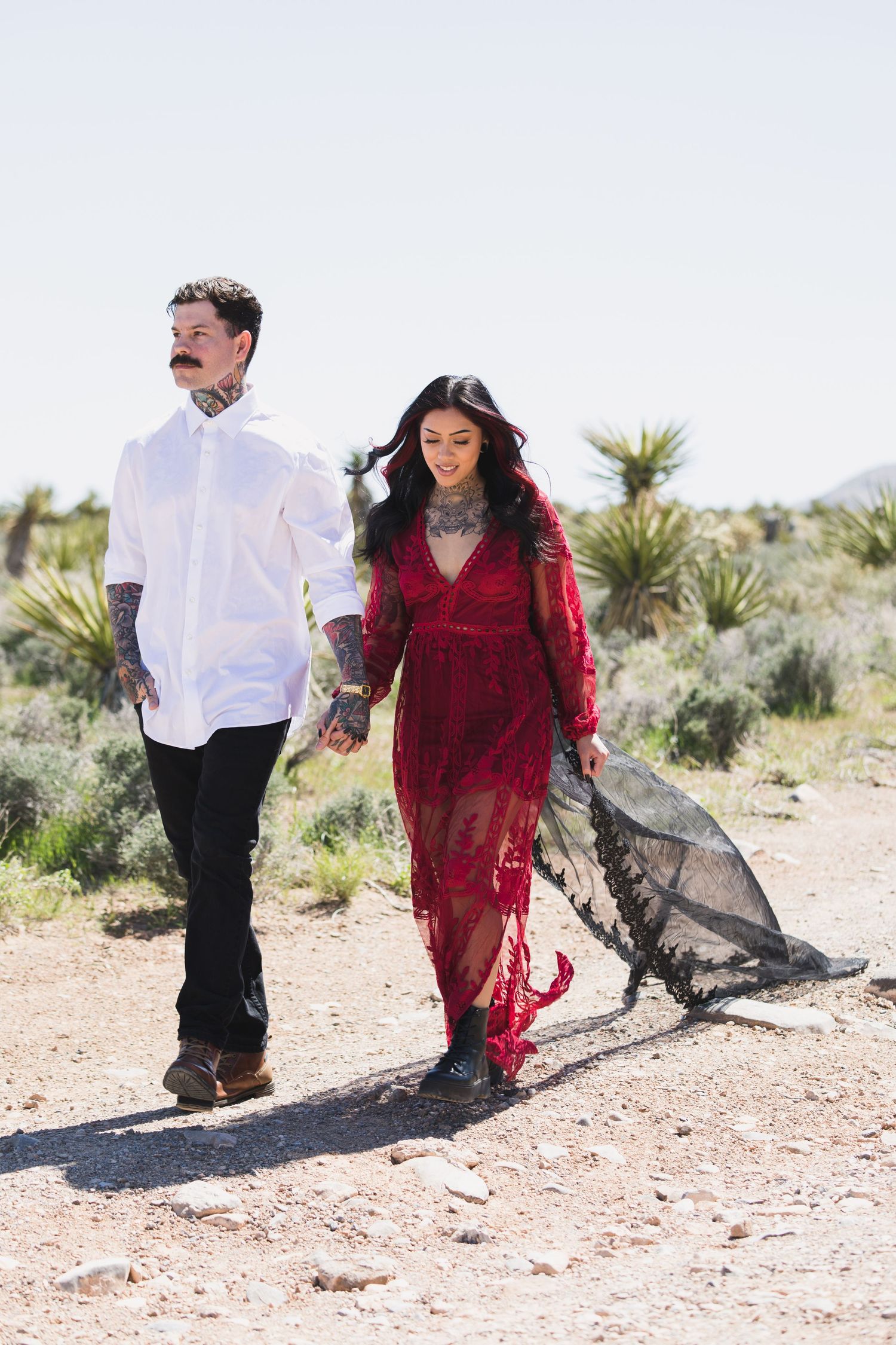 Couple walks hand in hand through desert landscape, wearing red dress and white shirt.