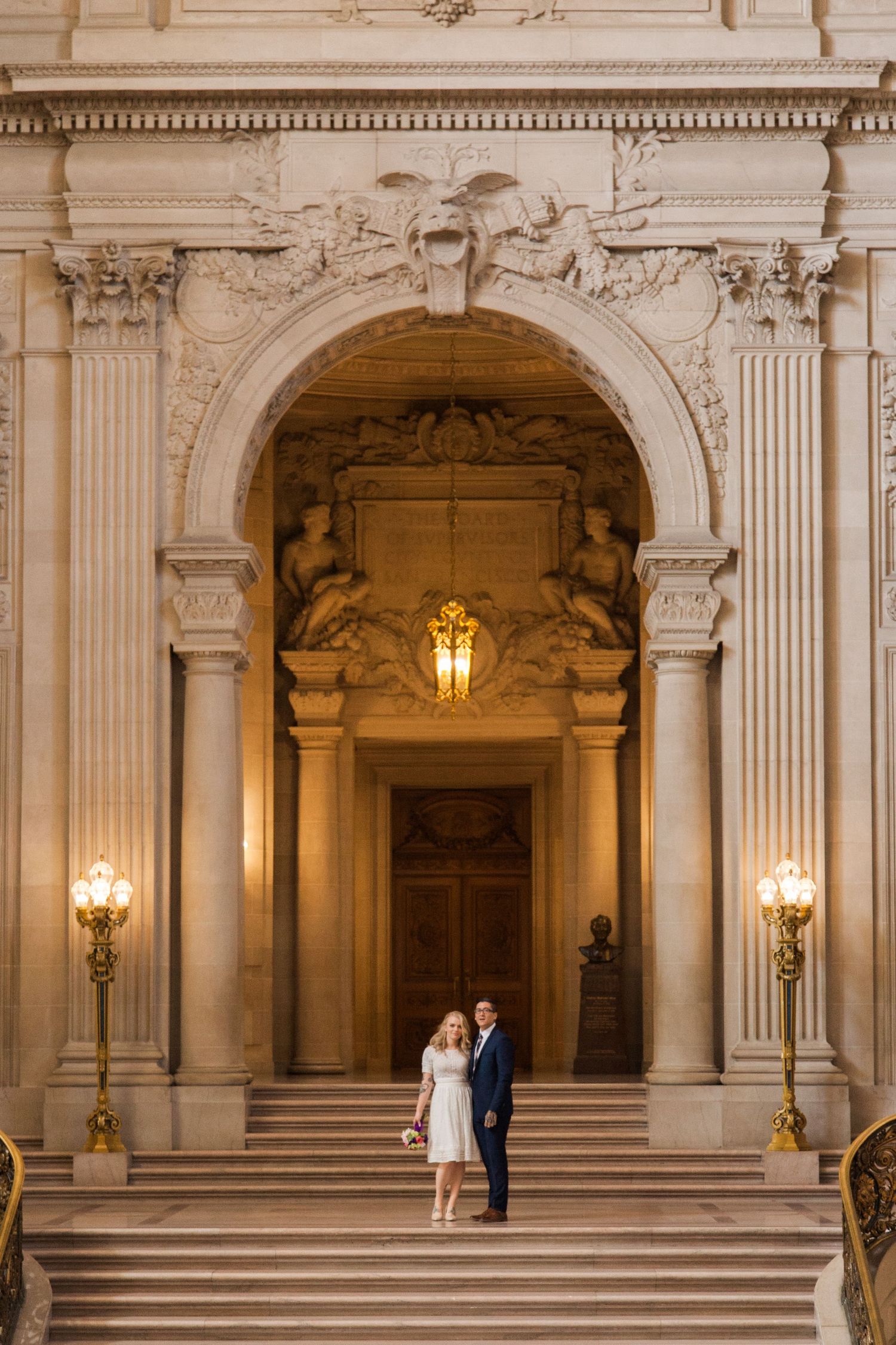 Couple standing on grand staircase beneath ornate classical architecture with warm lighting.