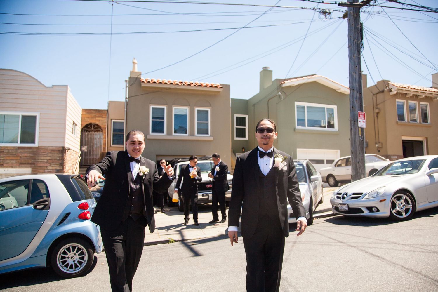 Groomsmen in formal black tuxedos walking down a residential street on wedding day.