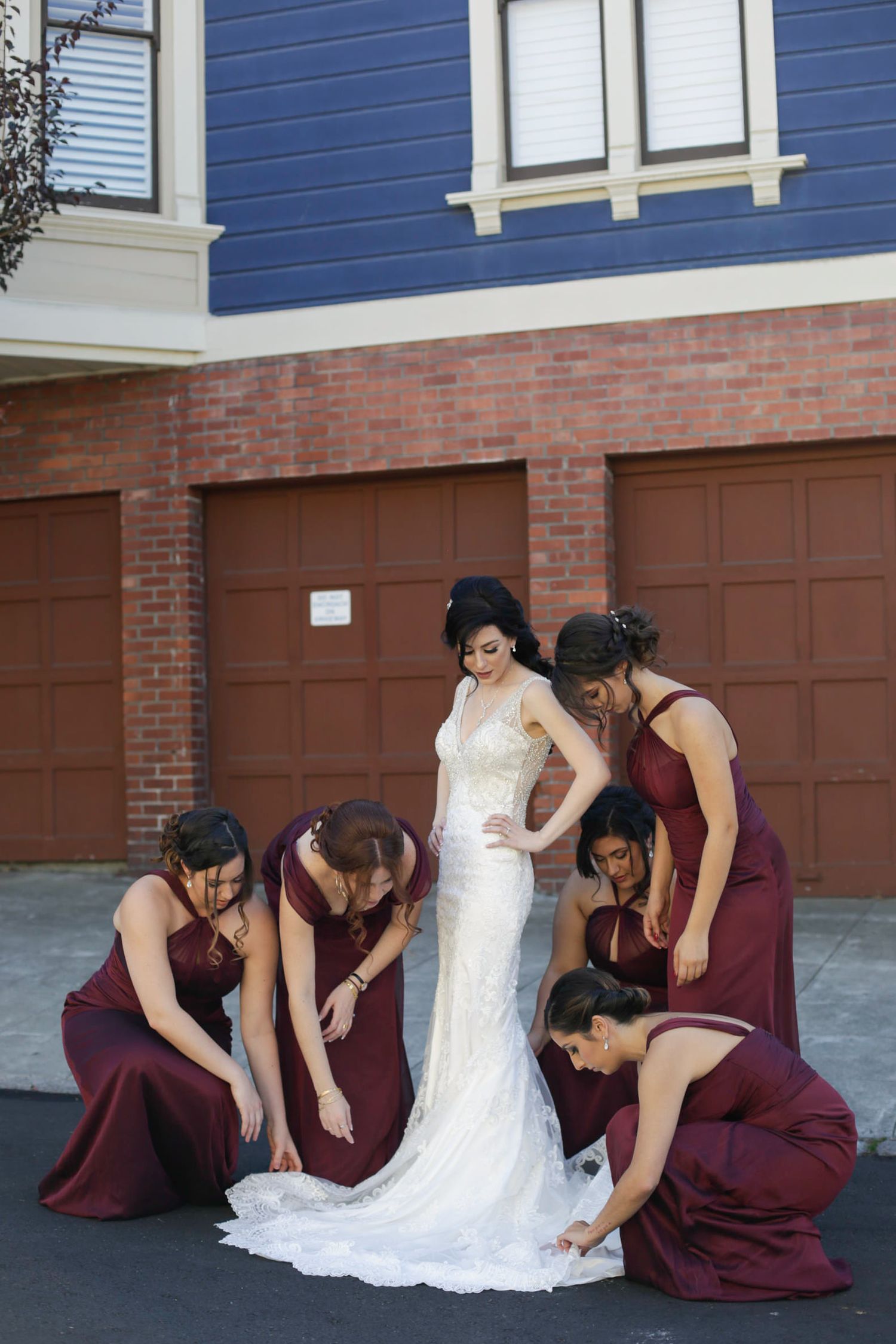 Bridesmaids in burgundy dresses help adjust the bride's wedding gown outdoors.