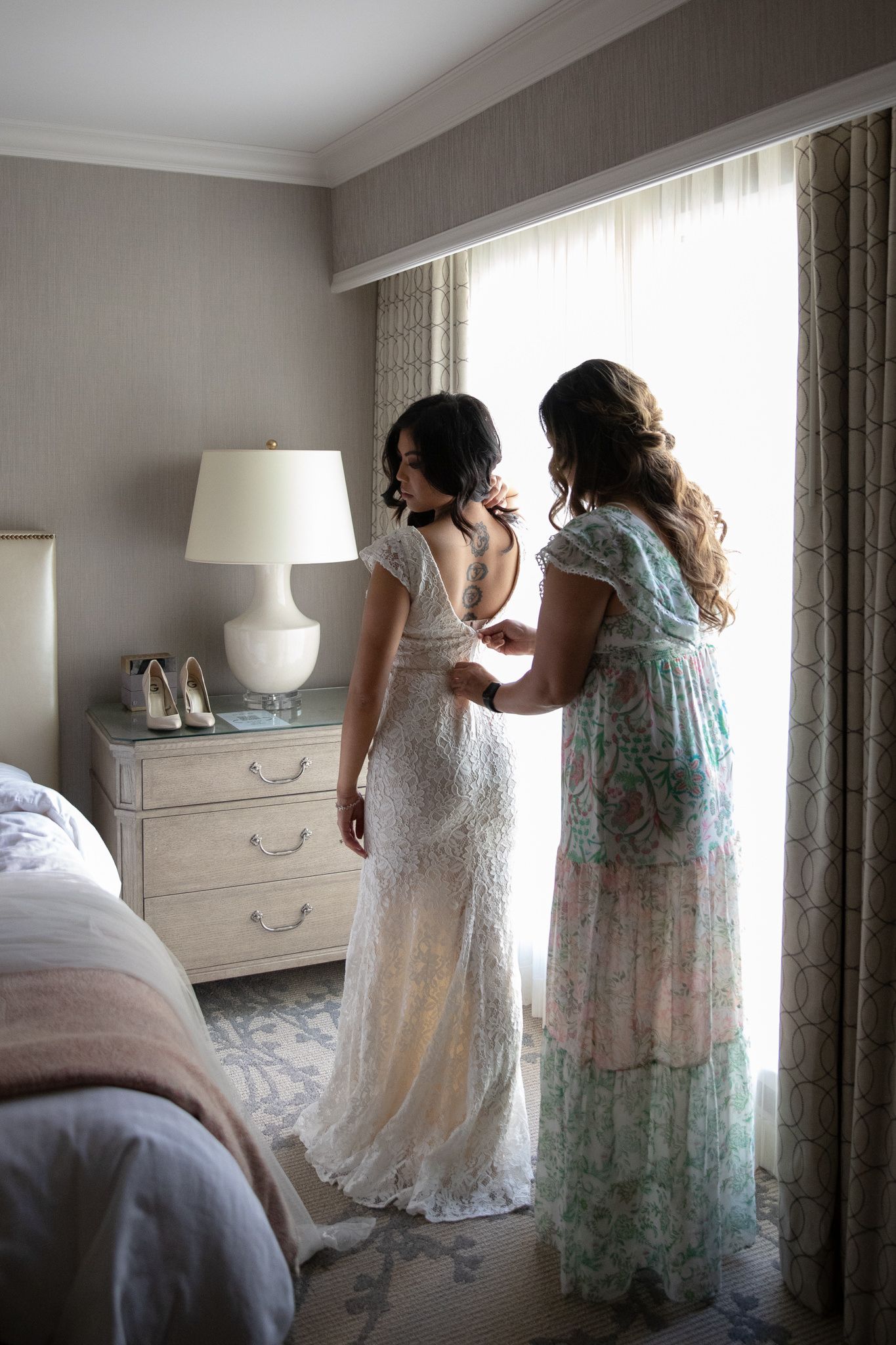 A tender moment of dress preparation as two women in white and floral dresses stand by a window in a bedroom.