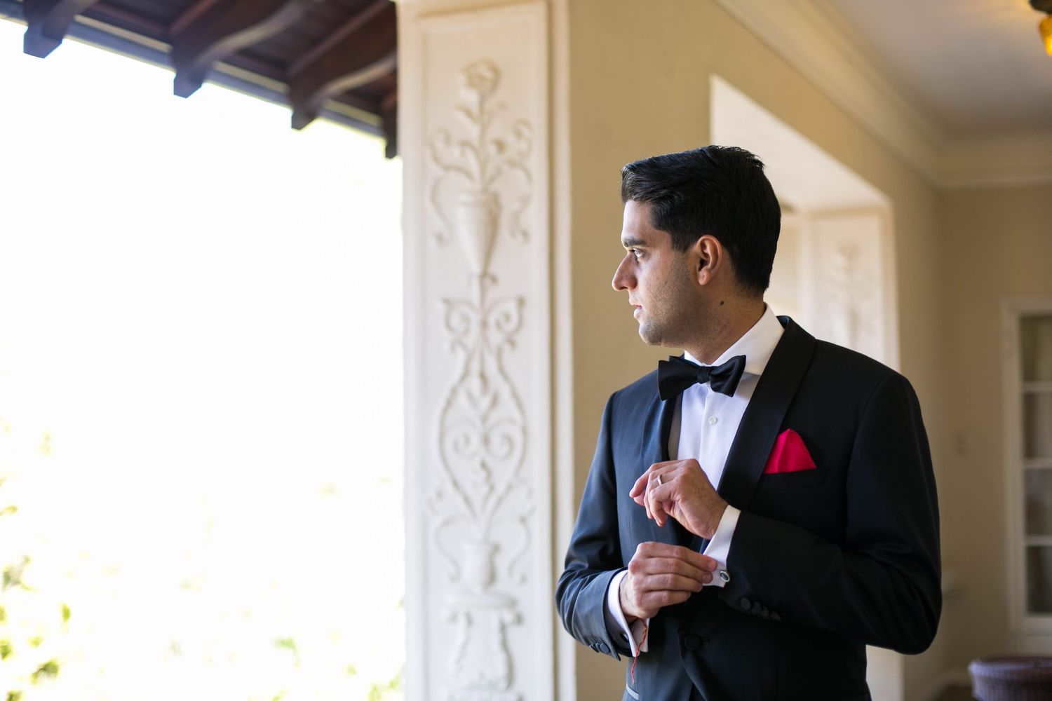 Elegant groom in black tuxedo with red pocket square adjusts his cuffs in a bright hallway.