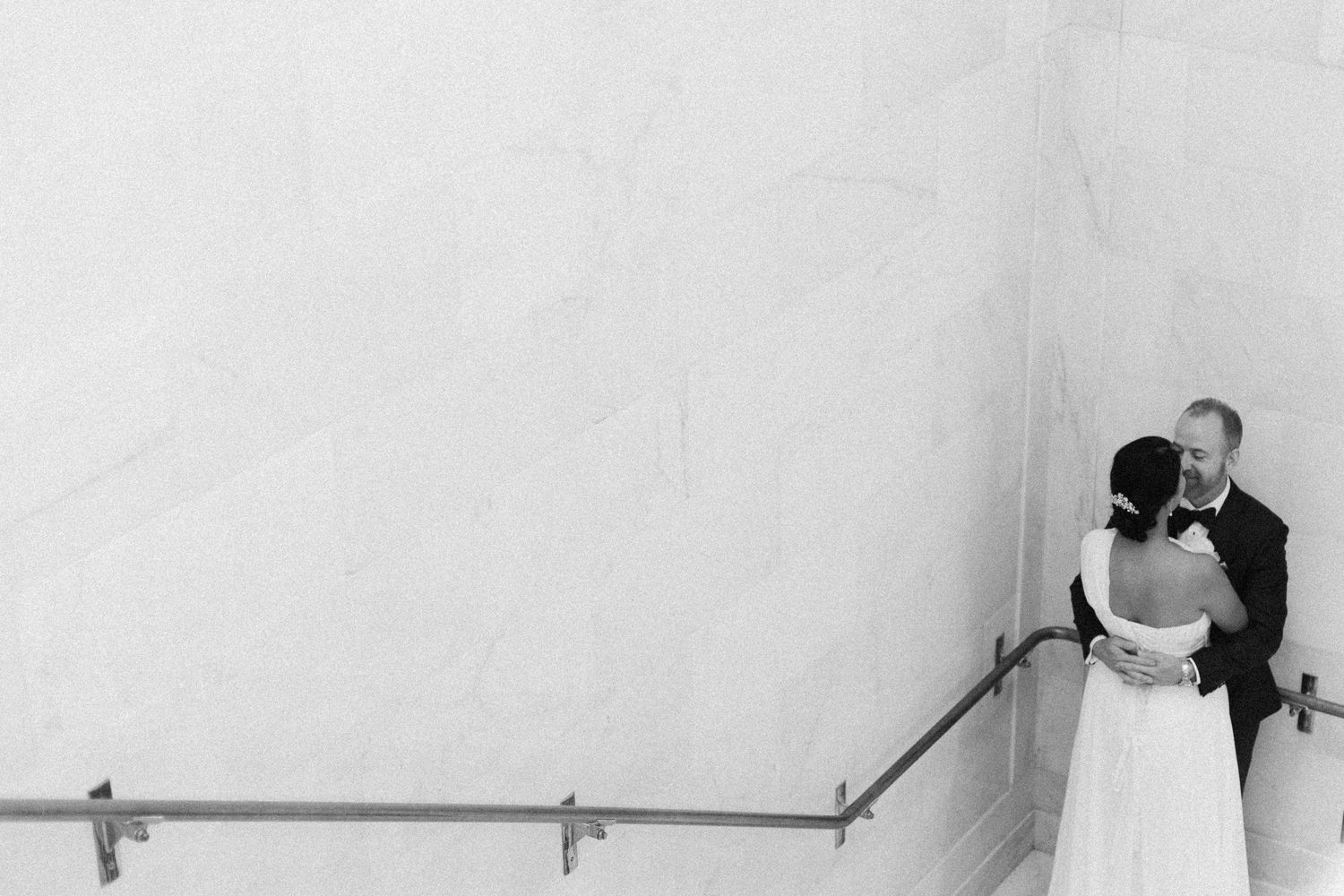 A minimalist black and white wedding portrait against a white wall with handrail.