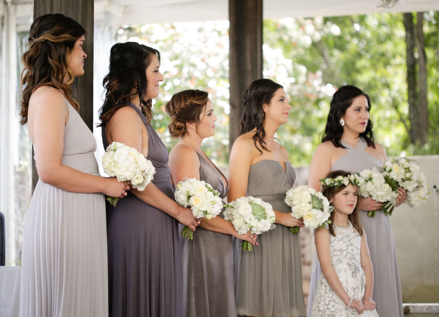 Row of bridesmaids in varying shades of gray dresses standing together at outdoor venue.