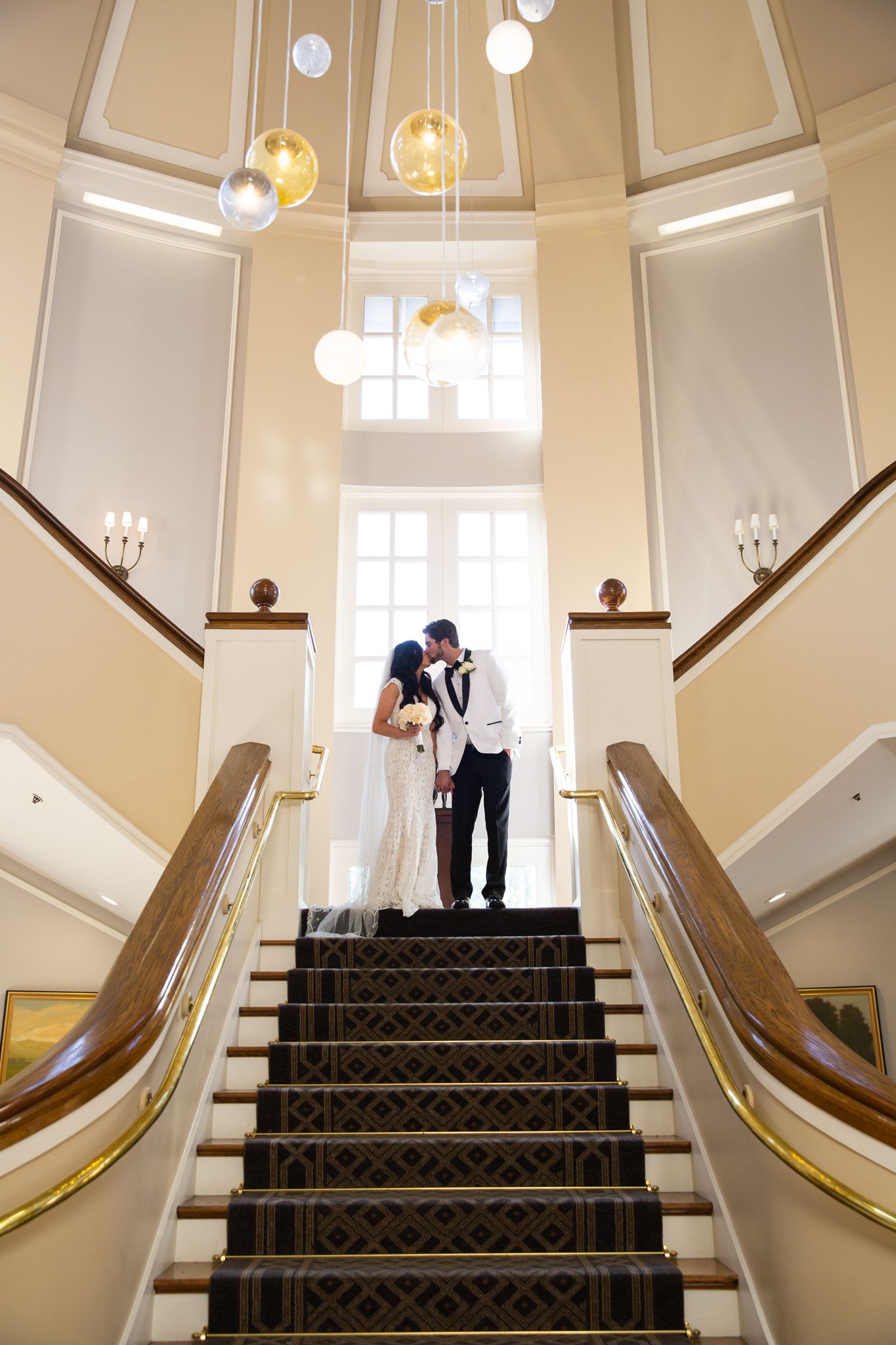Newlyweds share an intimate moment at the top of an elegant staircase with hanging pendant lights.