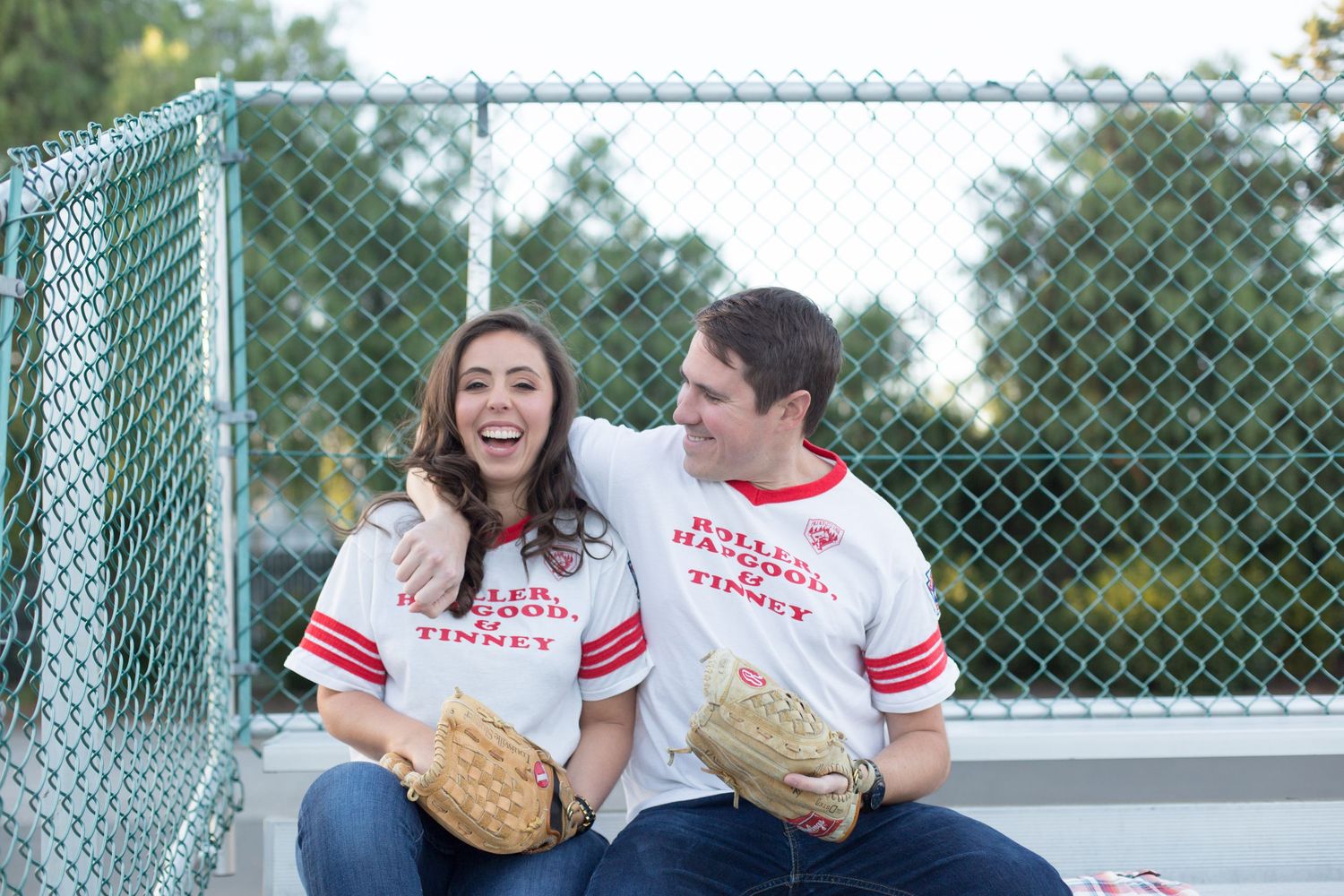 Couple in coordinated baseball jerseys laughing together against a chain-link fence.