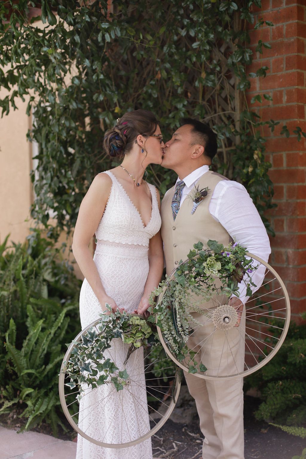 Wedding couple posing with vintage bicycle wheel against brick wall with ivy.