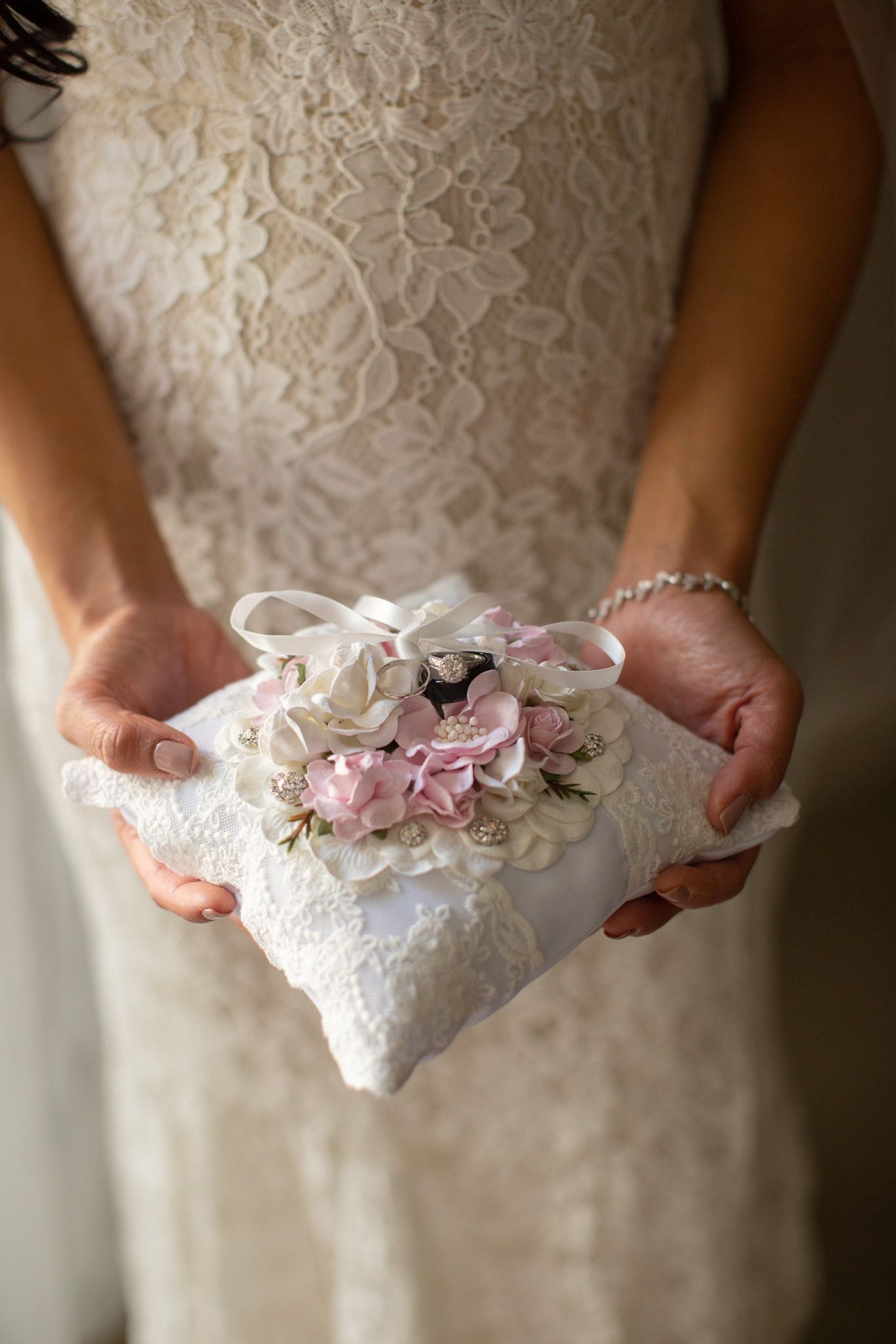 A delicate white lace wedding ring pillow held in hands against a wedding dress.