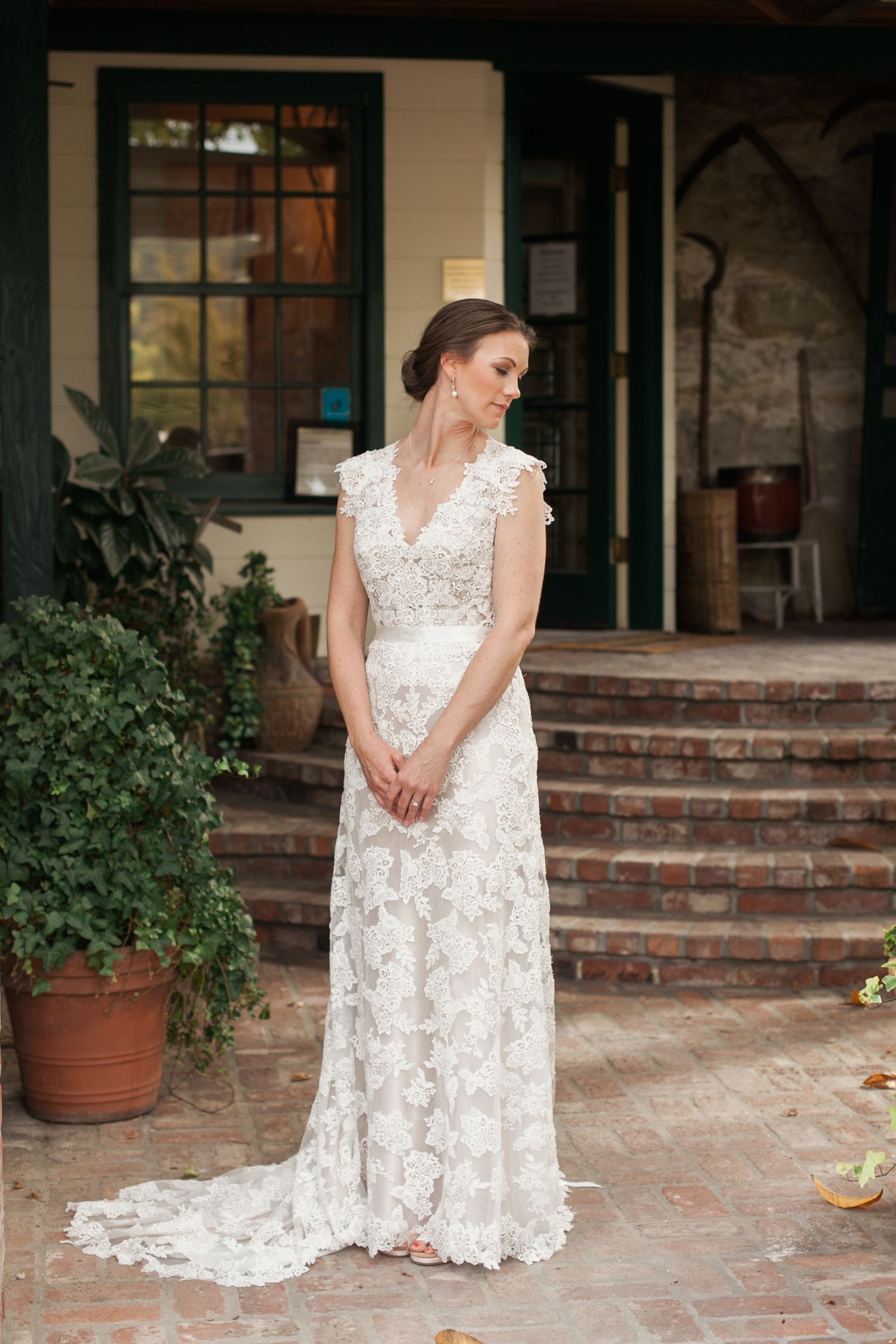 Bride in elegant lace wedding dress on brick patio with potted plants.