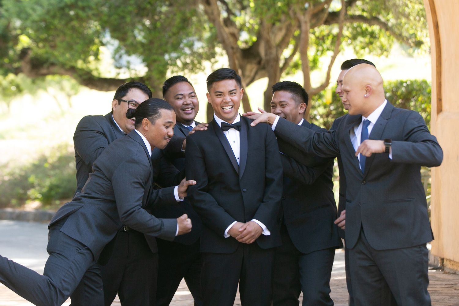 Groomsmen in black tuxedos share a playful moment outdoors.
