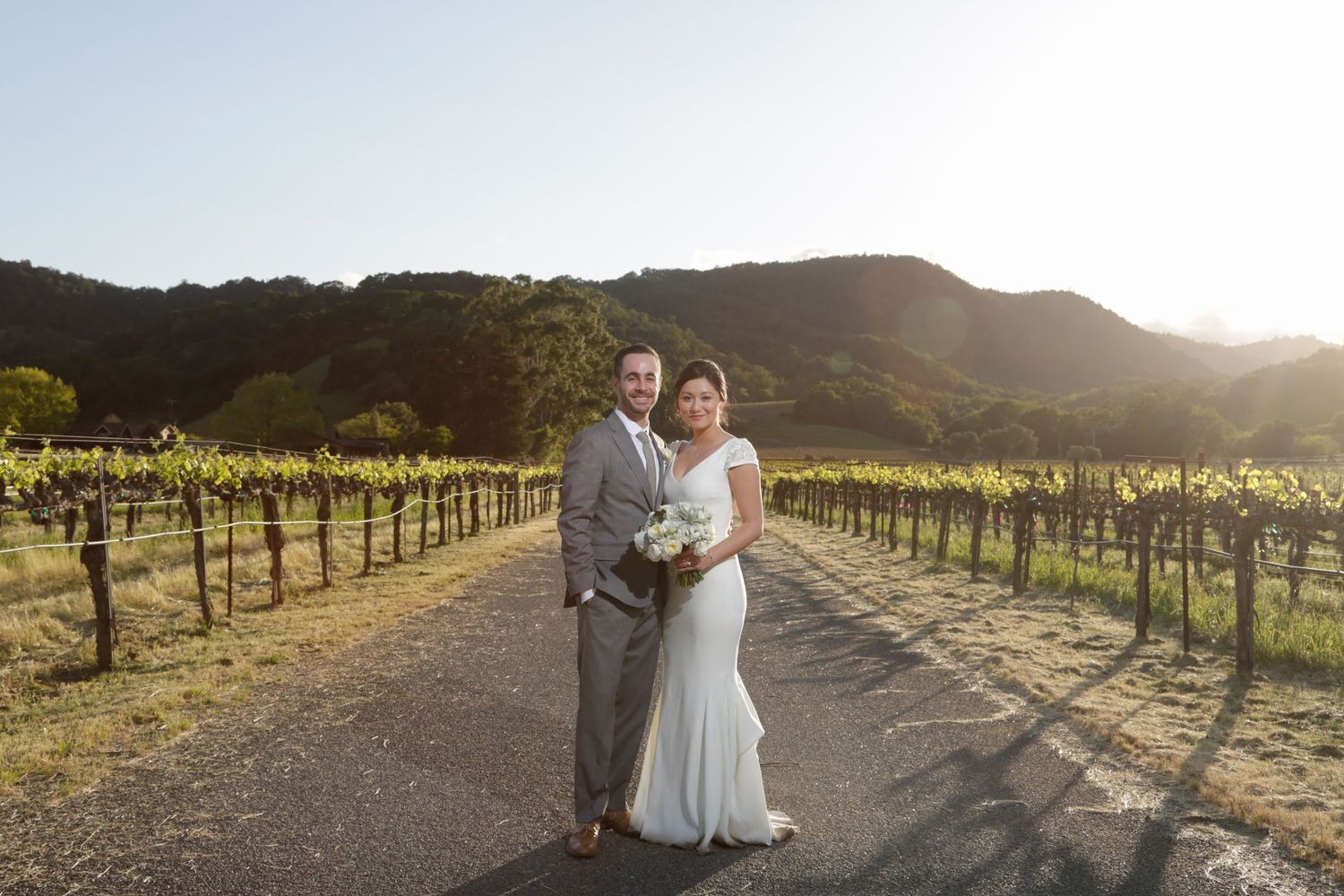 A couple poses together on a path between rows of grapevines at sunset.
