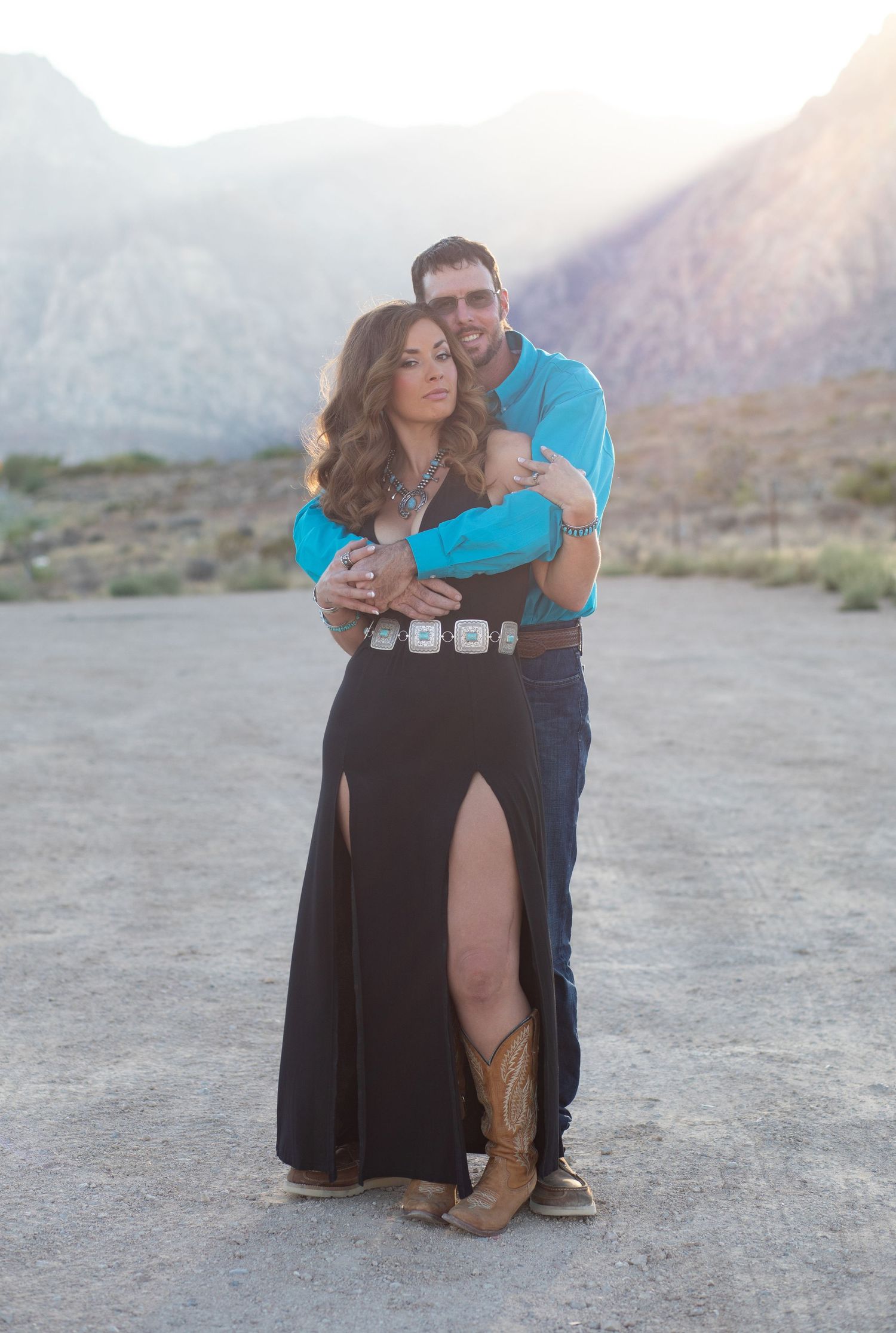 Romantic couple embraces in front of desert mountains, wearing western-inspired outfits with a long black dress and boots.