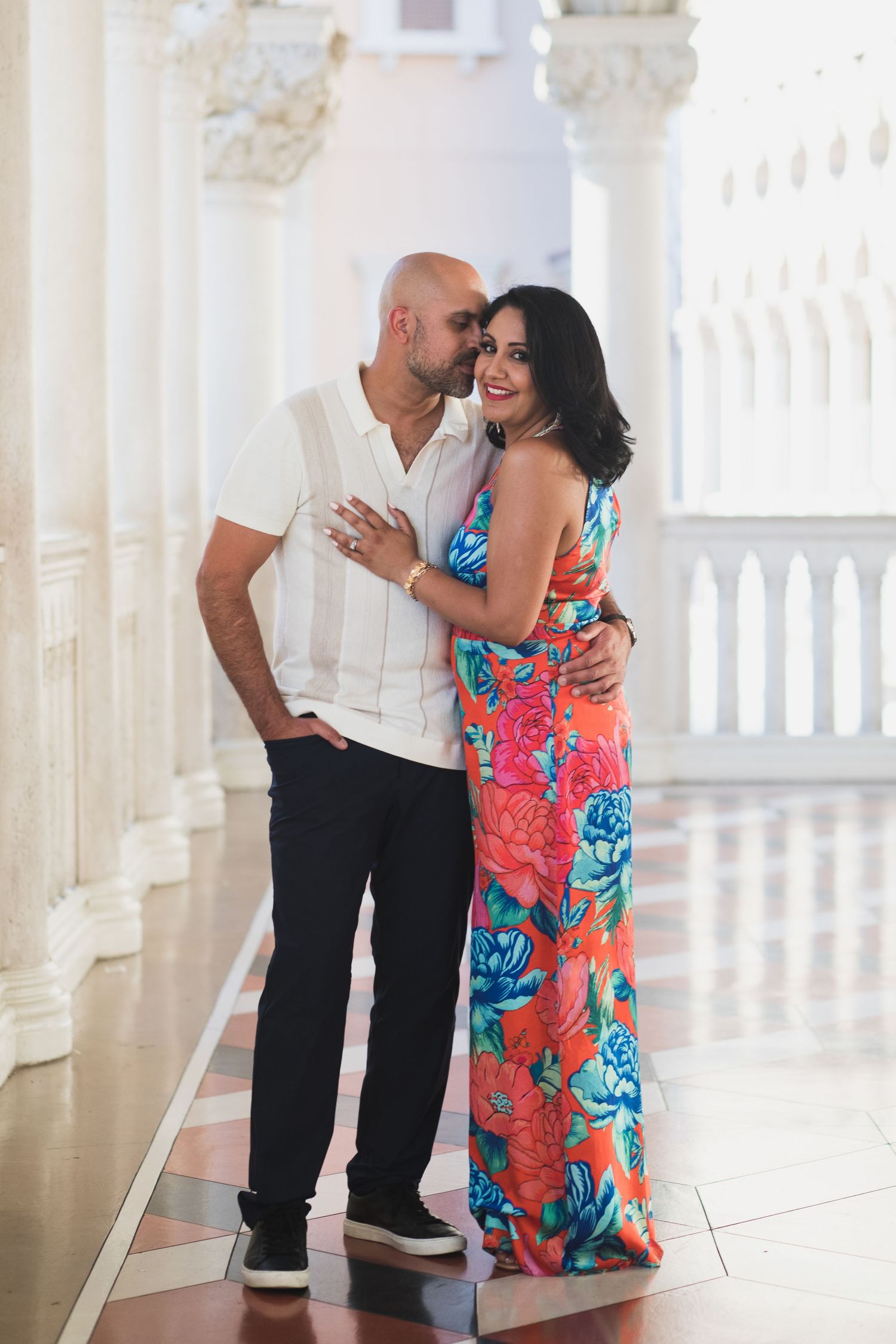 A couple embraces in a white columned hallway, with the woman wearing a vibrant floral maxi dress.