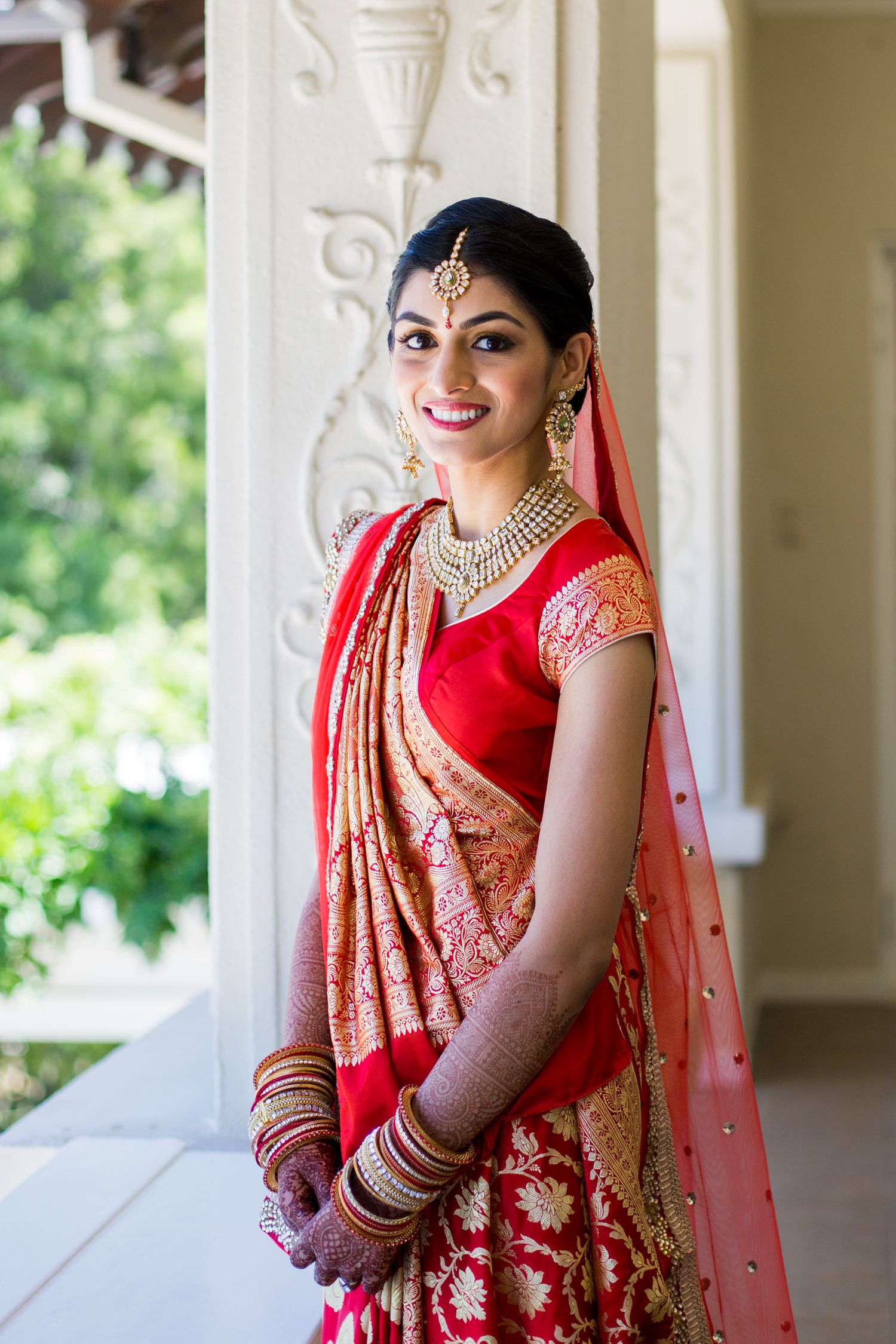 Traditional Indian bride in an ornate red lehenga posing gracefully in a columned veranda.