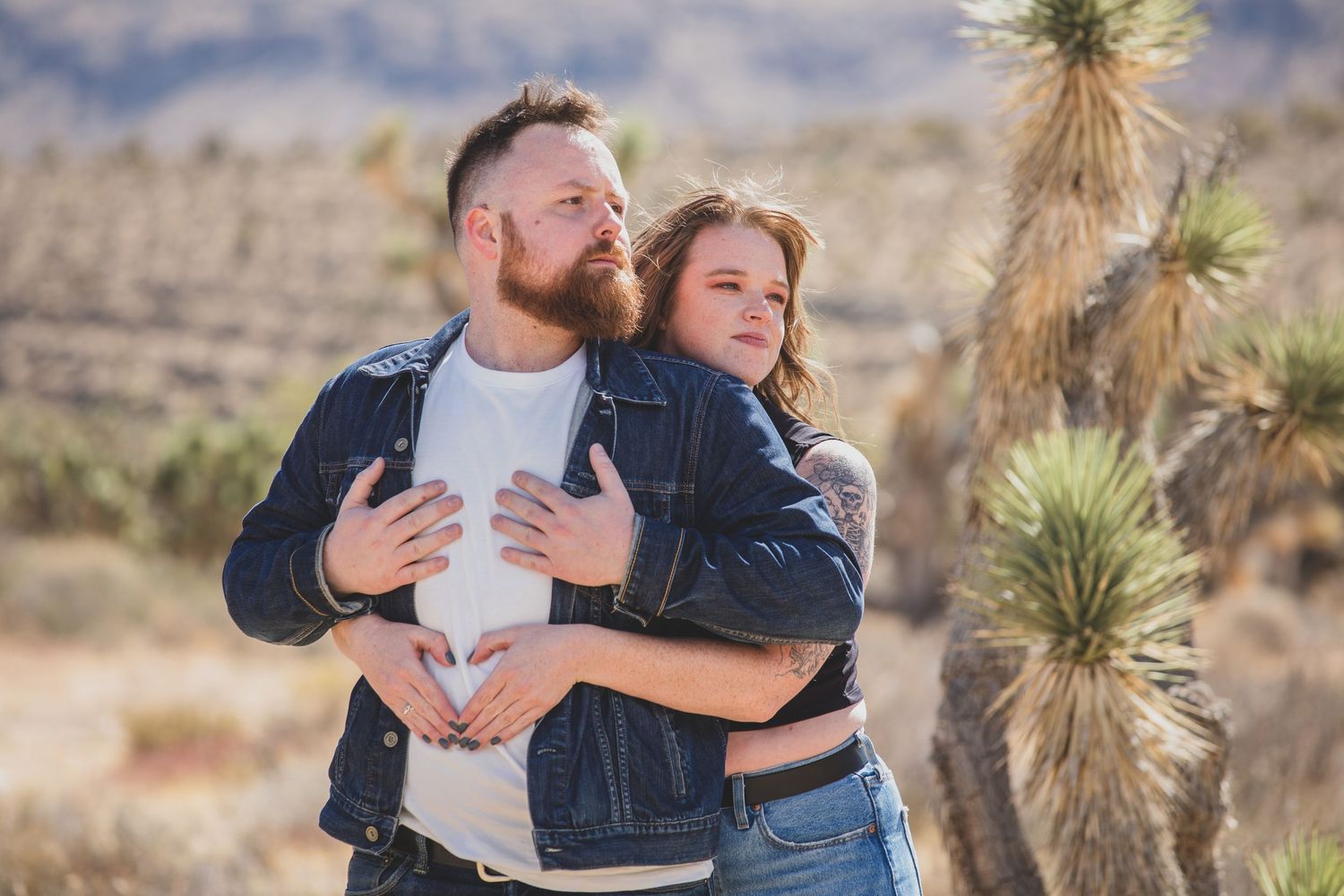 Romantic couple embraces in desert landscape with yucca plants and mountains in background.