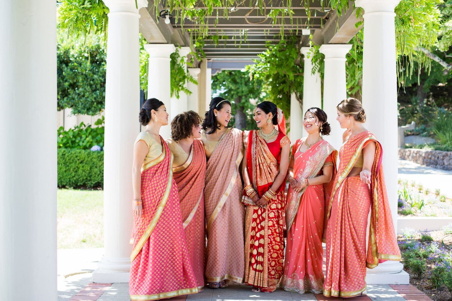 Group in coral and peach colored saris posing under columned garden walkway.