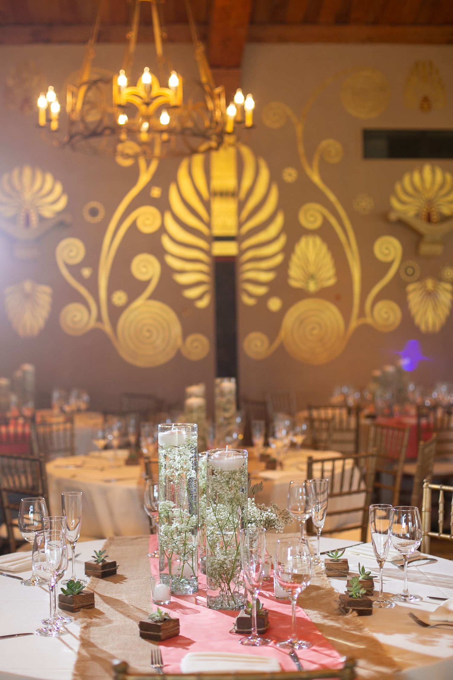 Wedding reception table settings with floral centerpieces against wall with decorative gold leaf patterns.