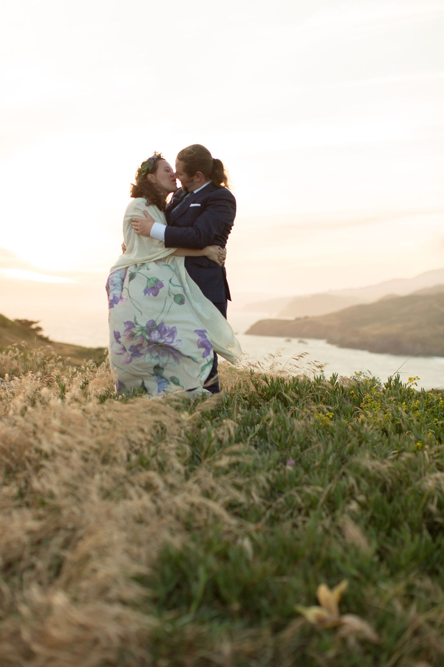 A romantic couple embracing on a hillside overlooking a scenic vista at sunset.