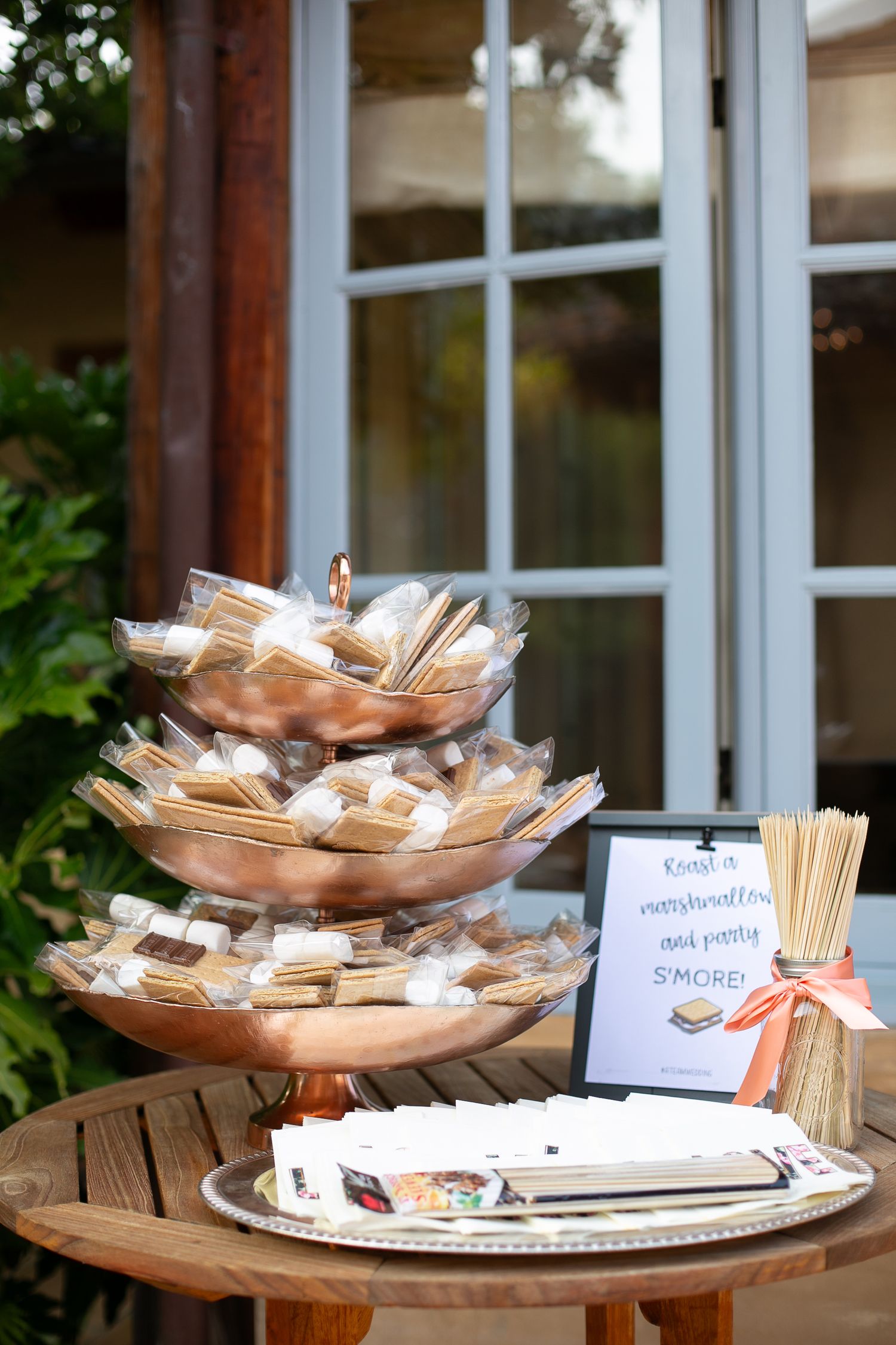 Tiered copper serving dishes with packaged wedding favors displayed on a wooden table by a window.