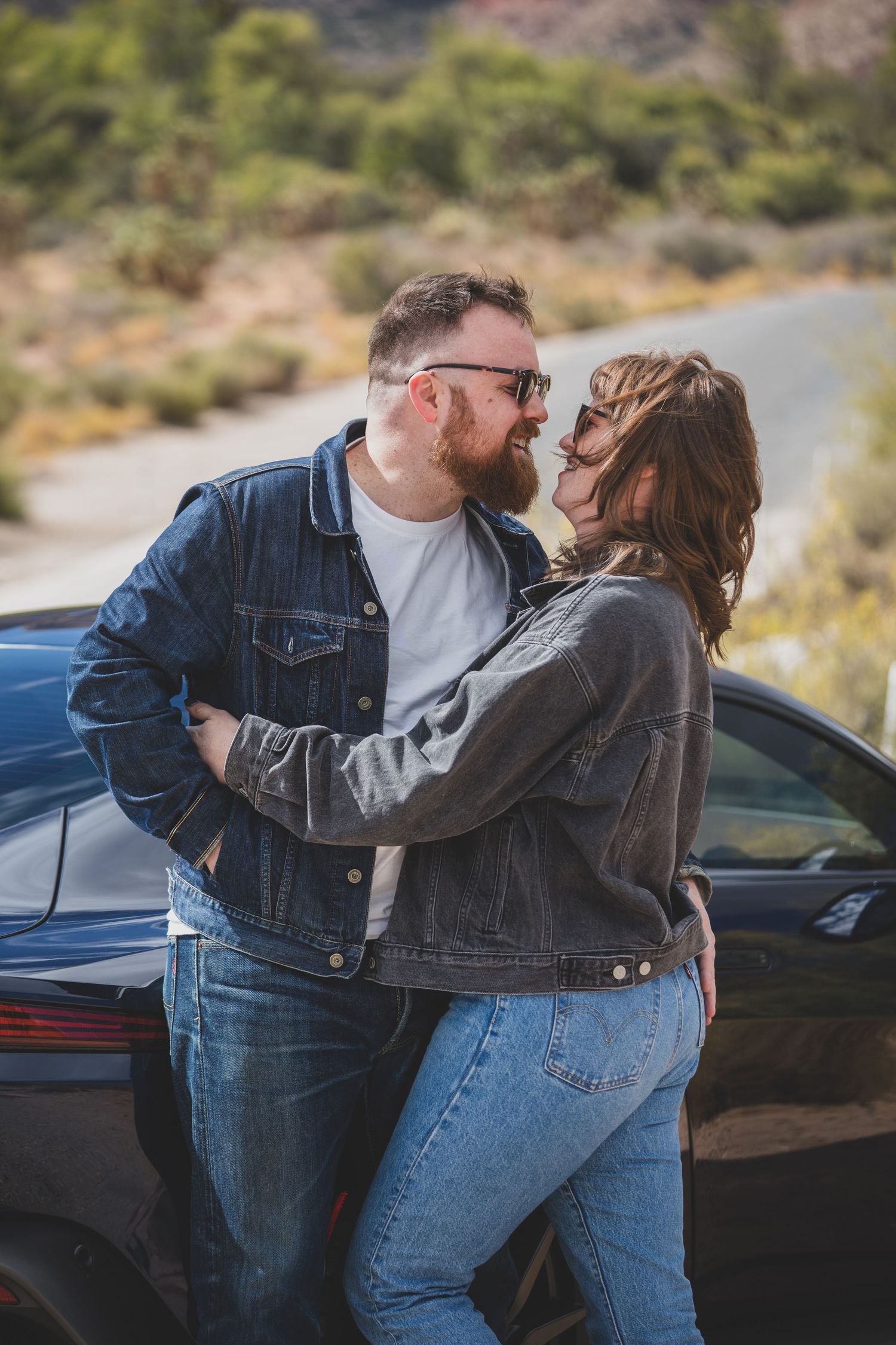 A couple in denim jackets embracing by their car on a desert road.