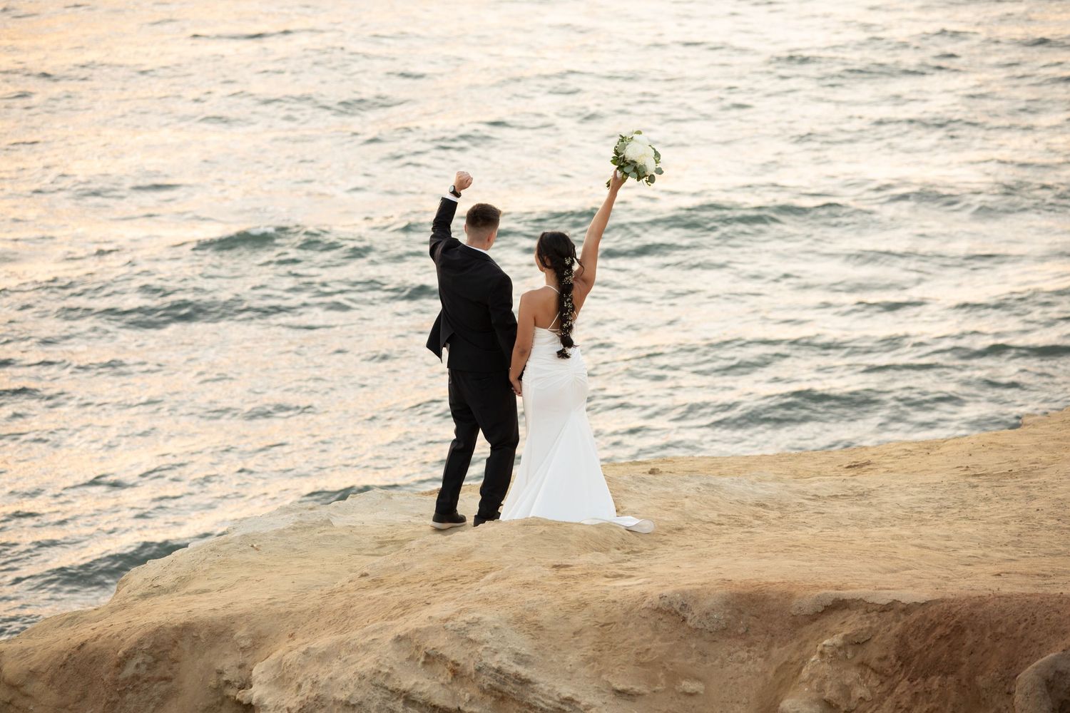 A celebratory pose by newlyweds on oceanside cliffs at sunset.