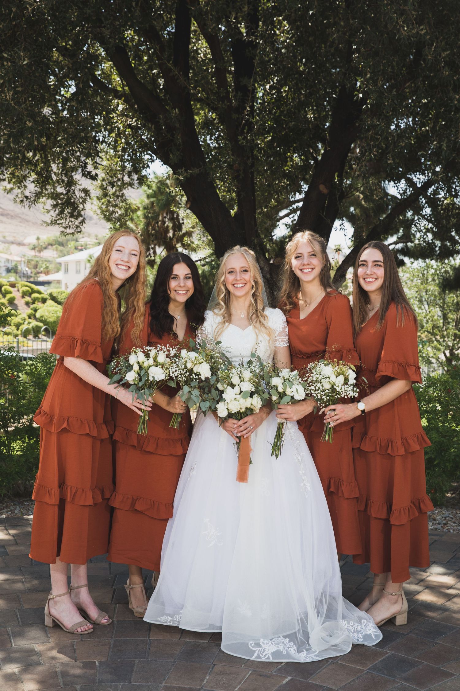 Bridal party in rust-colored dresses poses with bride in white gown outdoors.