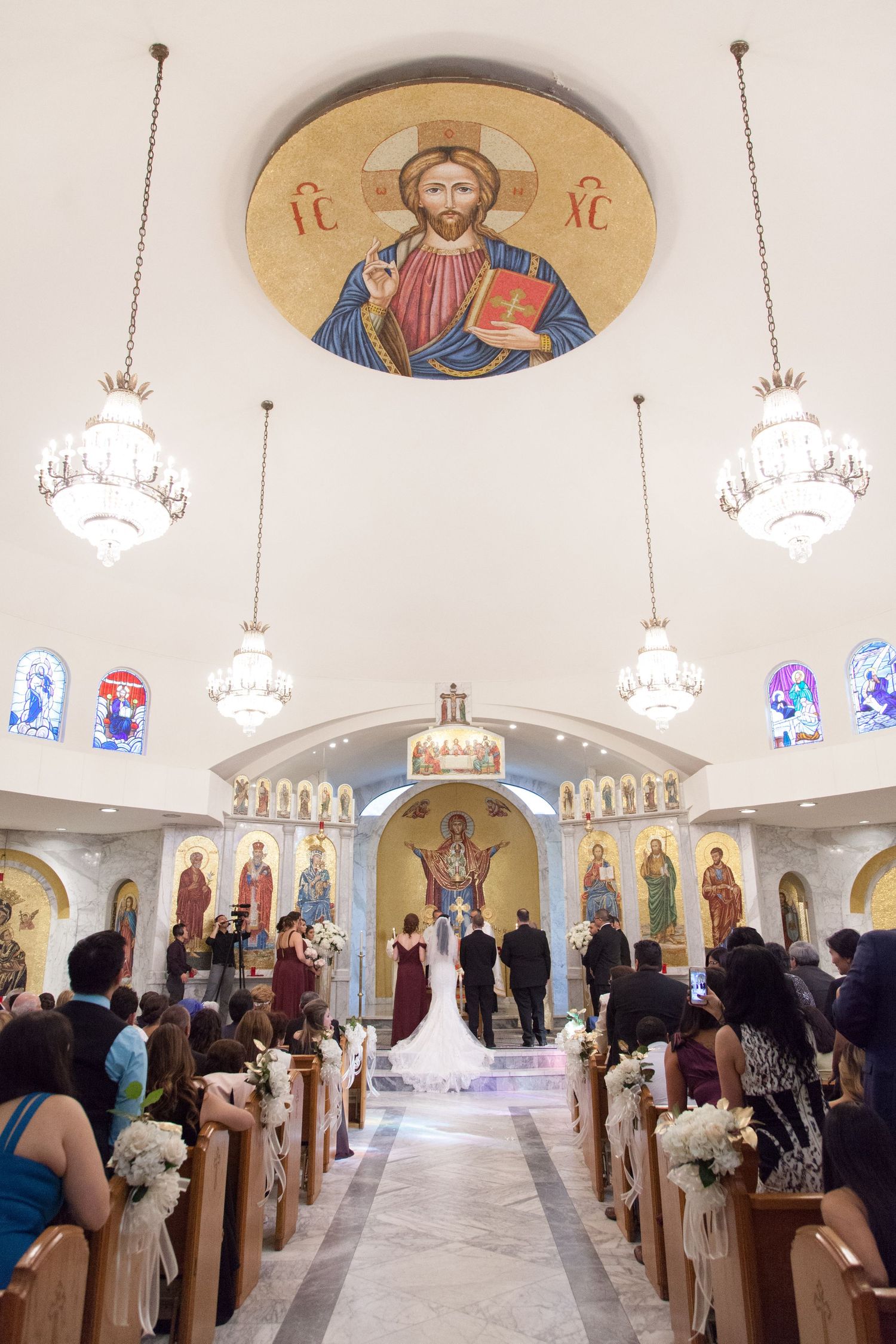 Orthodox church wedding ceremony with ornate religious artwork and crystal chandeliers.