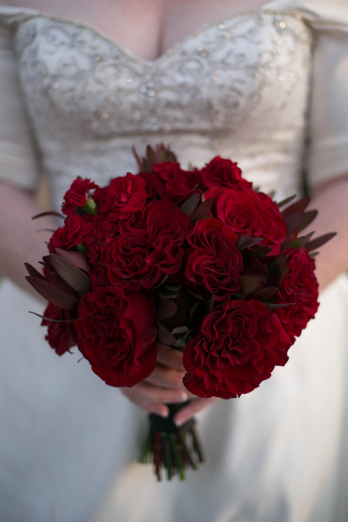 A bridal bouquet of deep red roses held against a white lace wedding dress.