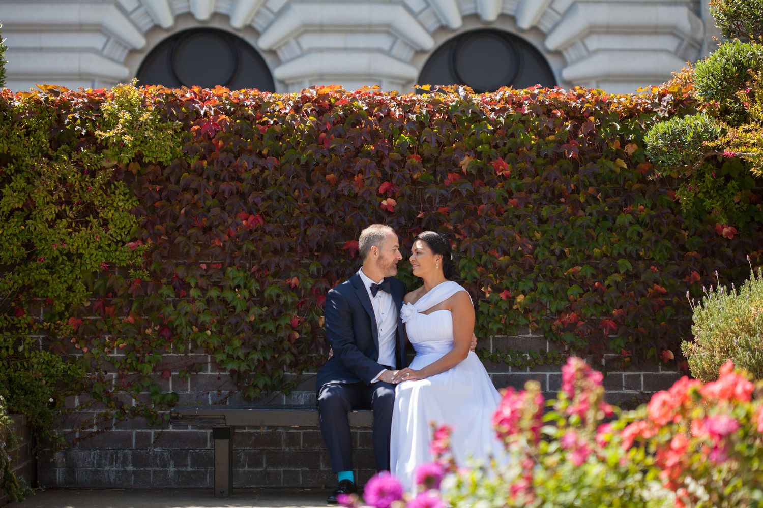 A wedding couple posing in front of a beautiful ivy-covered wall with blooming flowers.