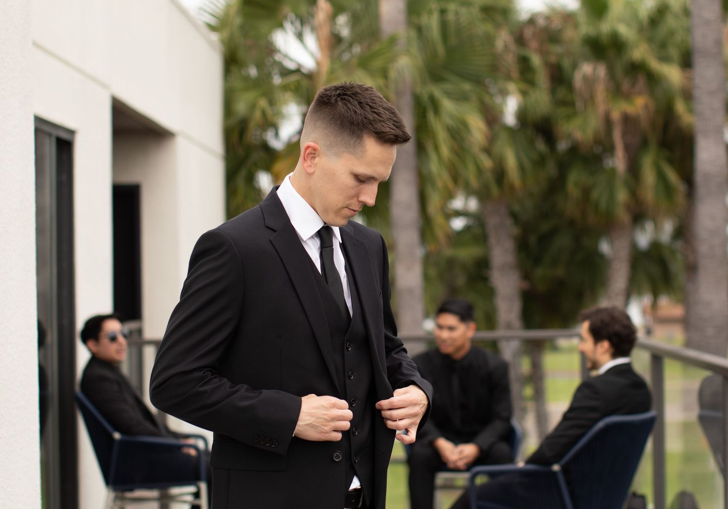 A person in a black suit adjusts their tie while standing outside.