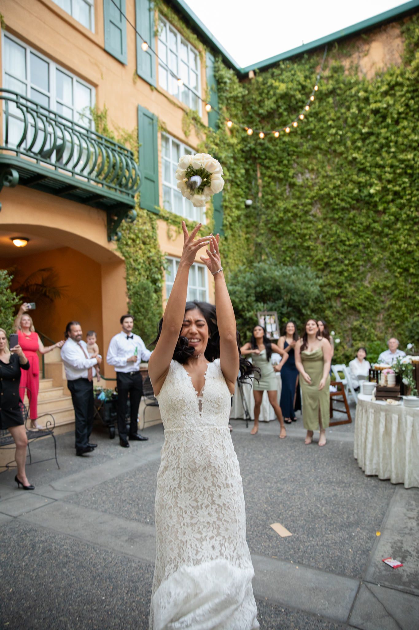 A bride in a lace gown tosses her bouquet during an outdoor wedding reception.