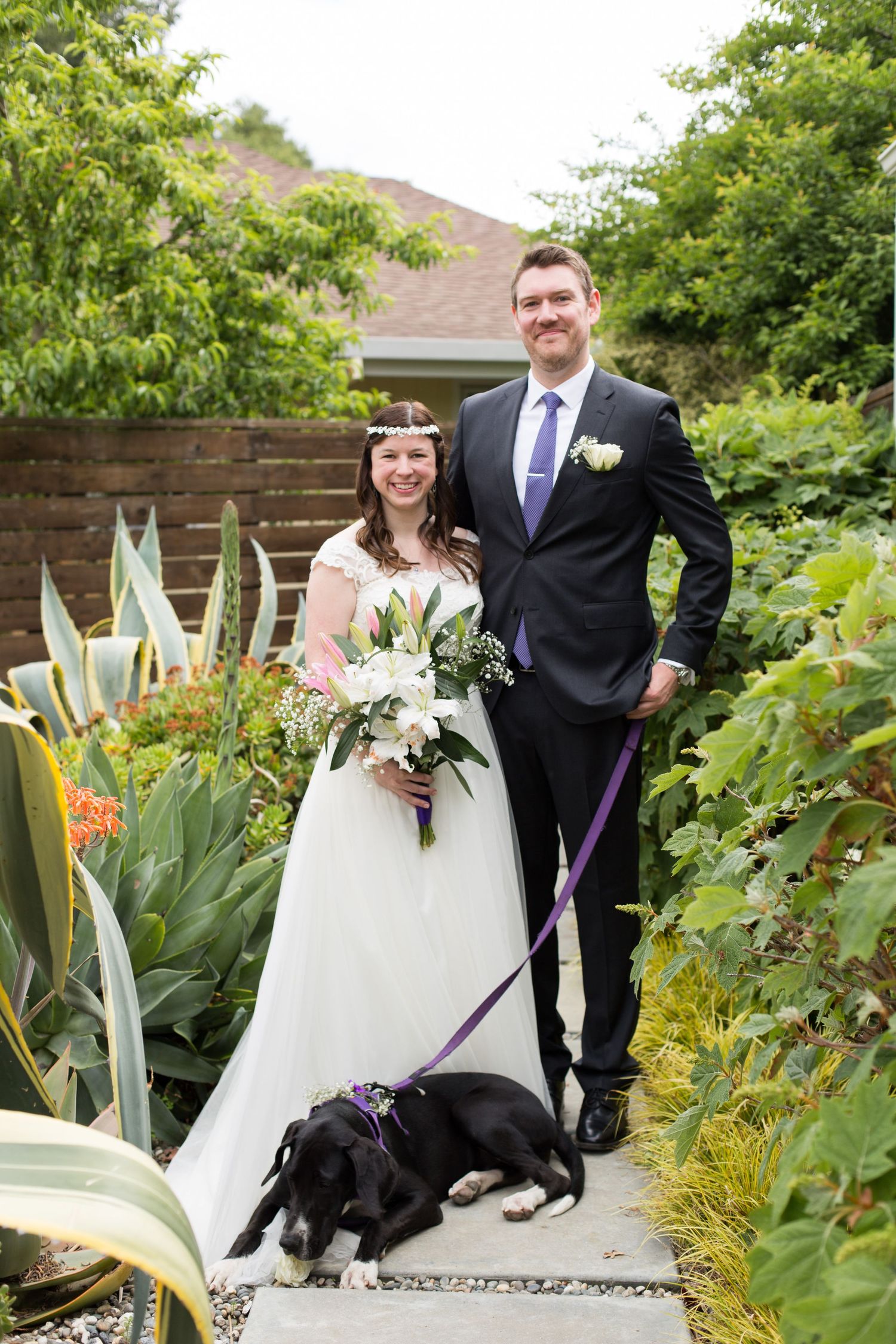Newlyweds pose in garden with their black dog wearing a purple leash.