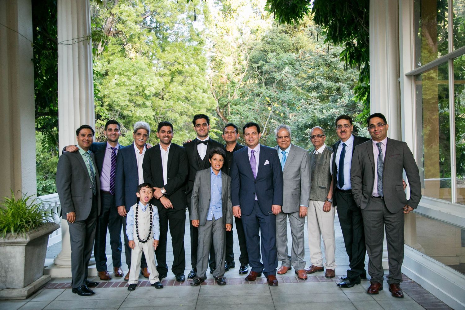 Group of formally dressed men pose together for a wedding party photo on an outdoor terrace.