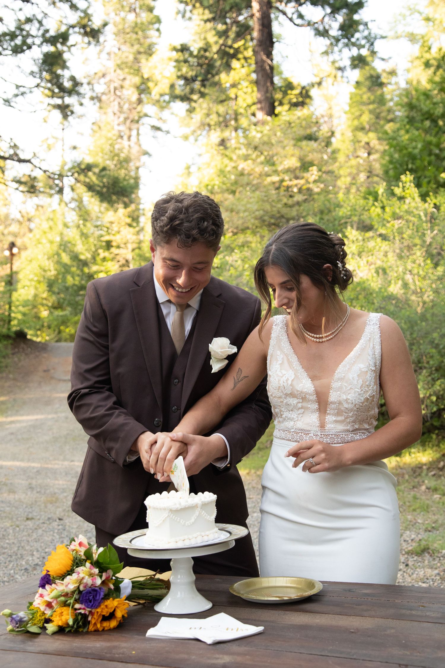 Couple in wedding attire cutting their white wedding cake together at an outdoor reception with floral decorations.