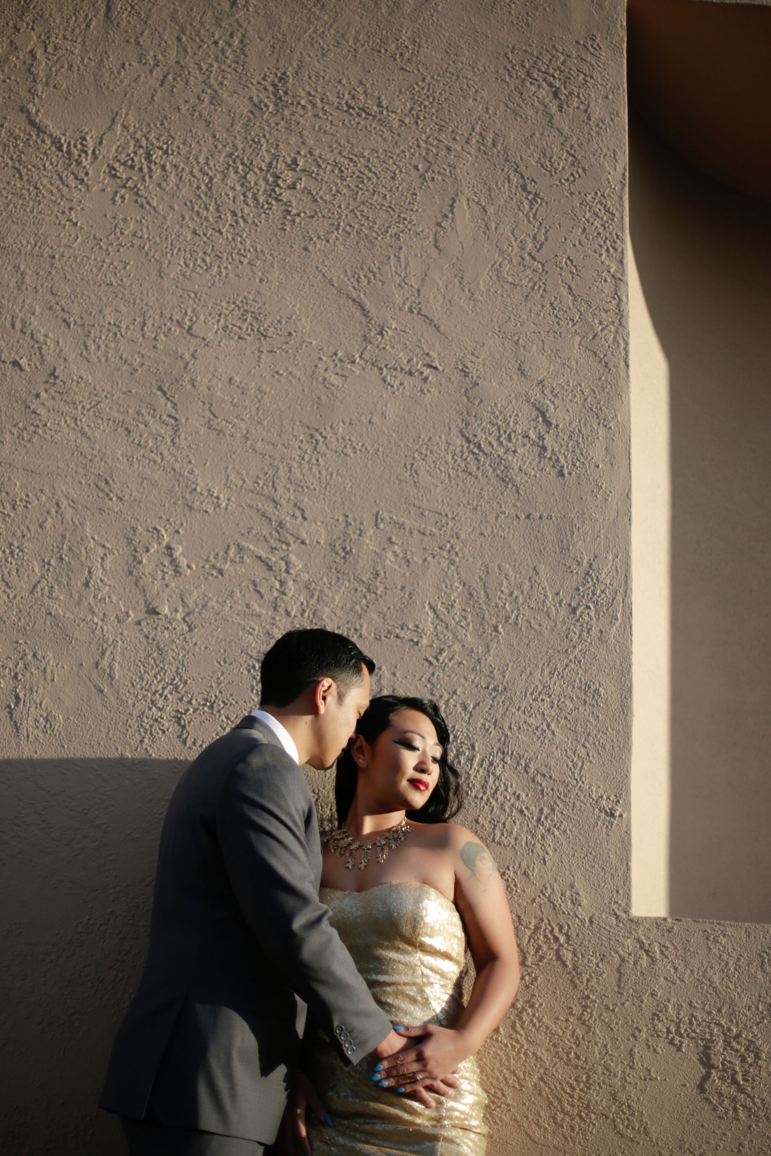 A couple in formal wear pose against a textured beige wall in dramatic lighting.