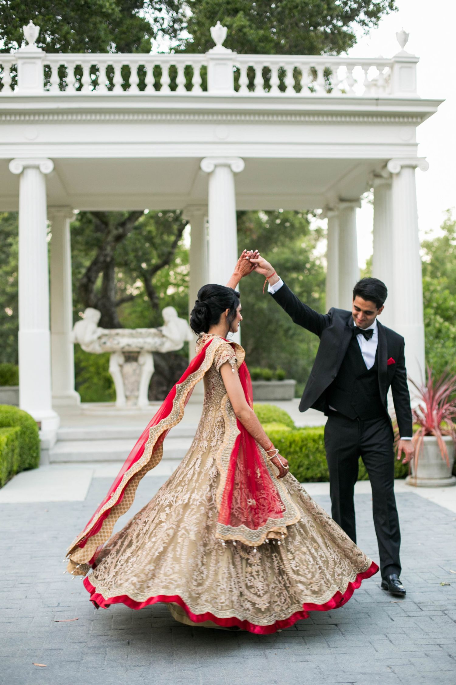 Elegant wedding attire with golden lehenga and red dupatta twirling in front of white pillared gazebo.