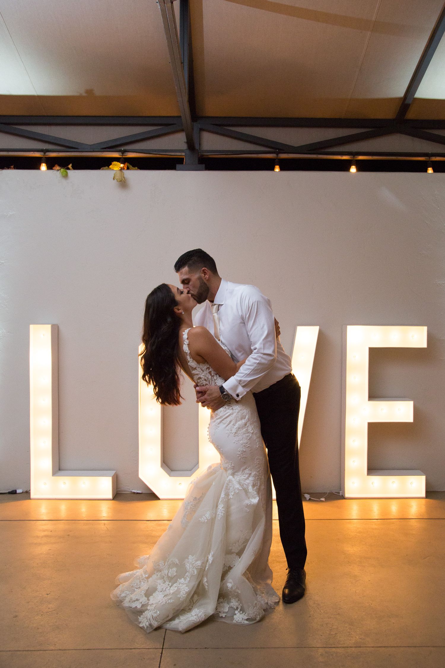 Backlit silhouette of couple sharing romantic moment in front of illuminated LOVE sign at wedding reception.