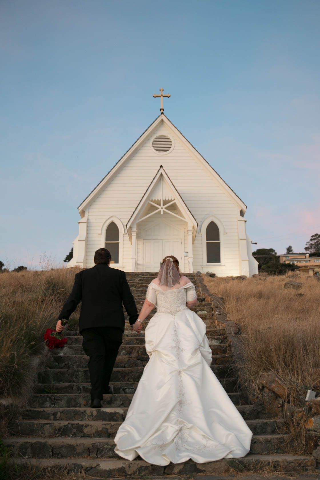 White chapel on a hillside with bride and groom ascending stone steps at sunset.