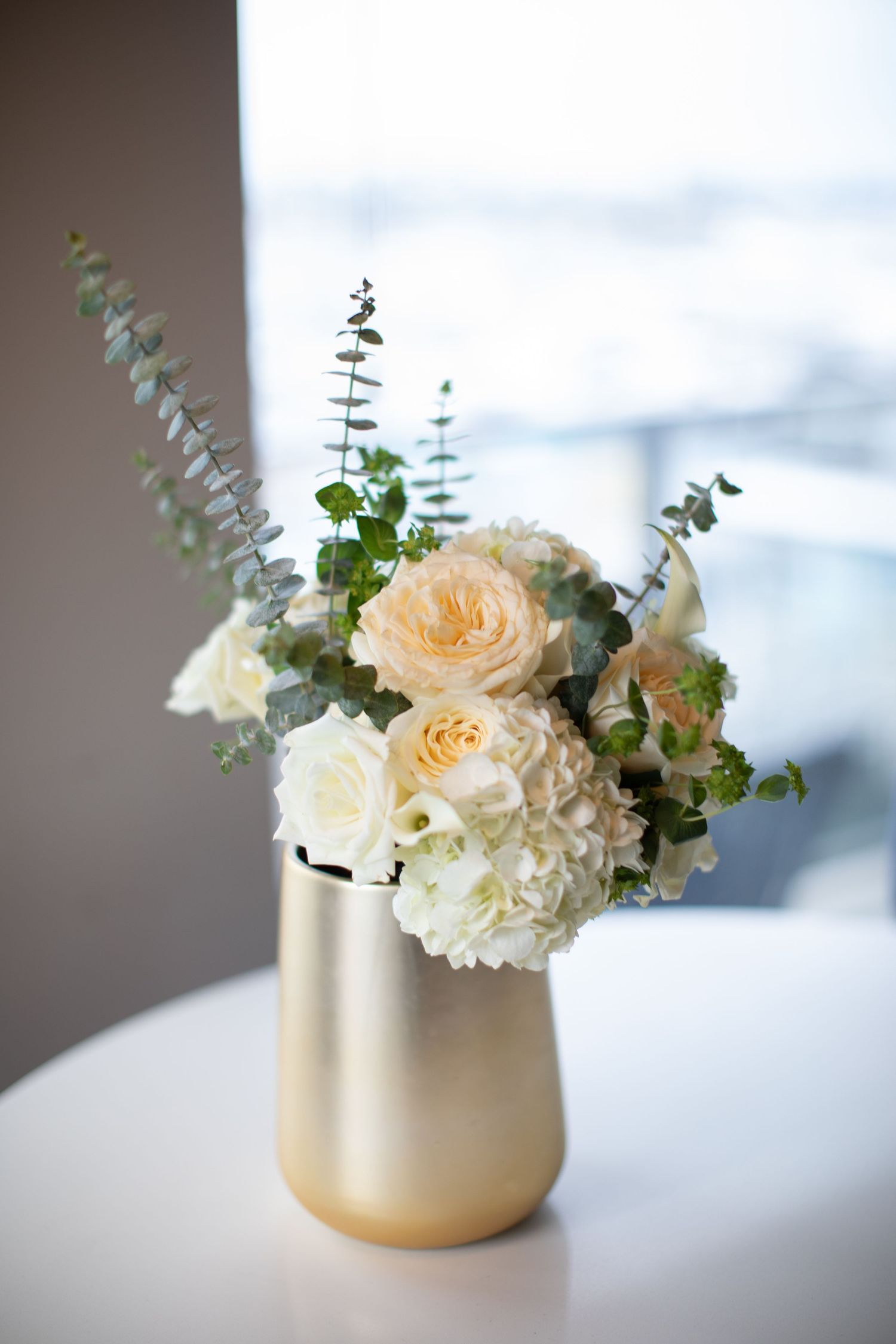 An elegant floral arrangement of peach and white roses with eucalyptus in a frosted gold vase.