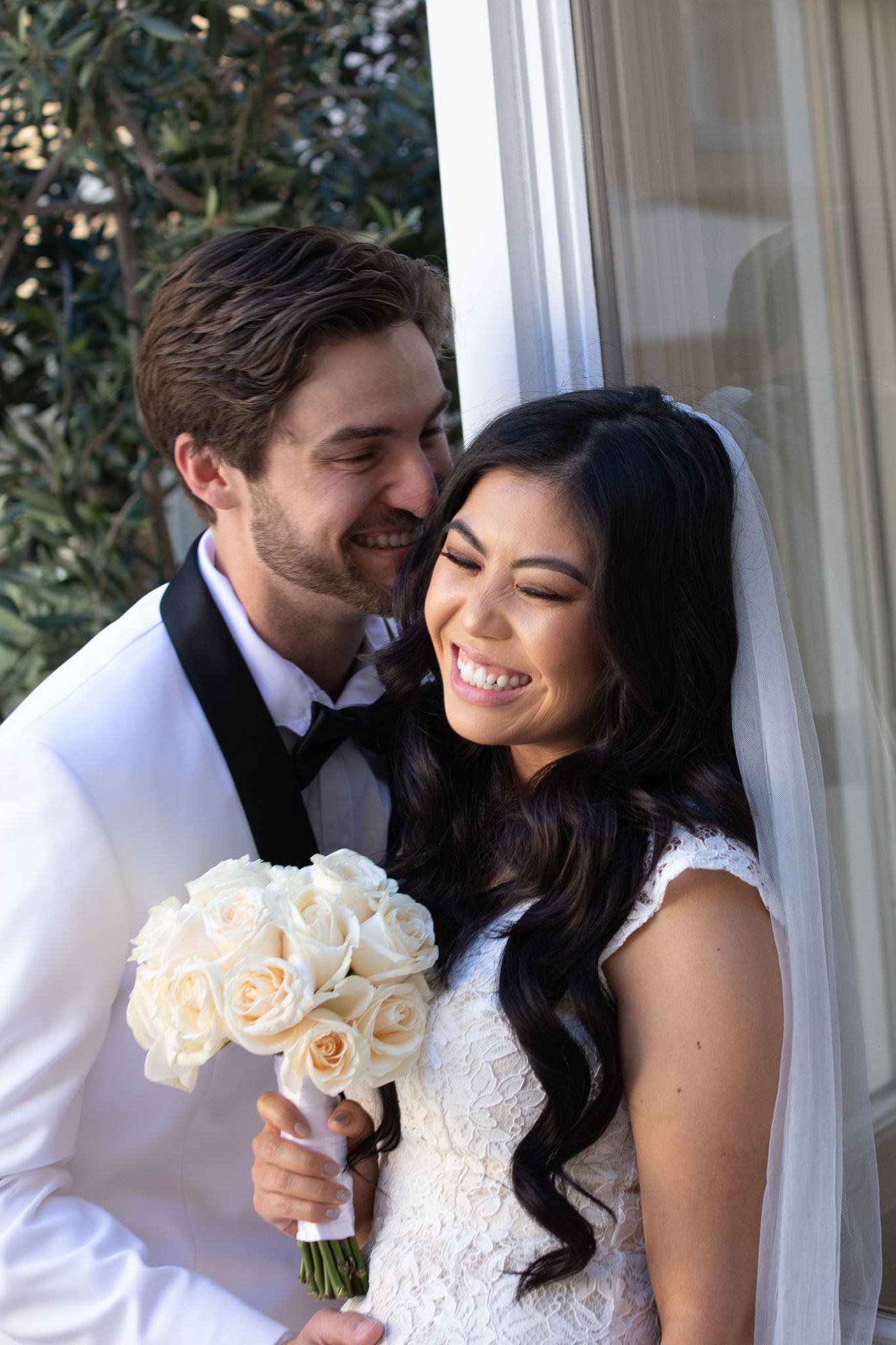 A joyful wedding couple embracing while holding a bouquet of white roses.
