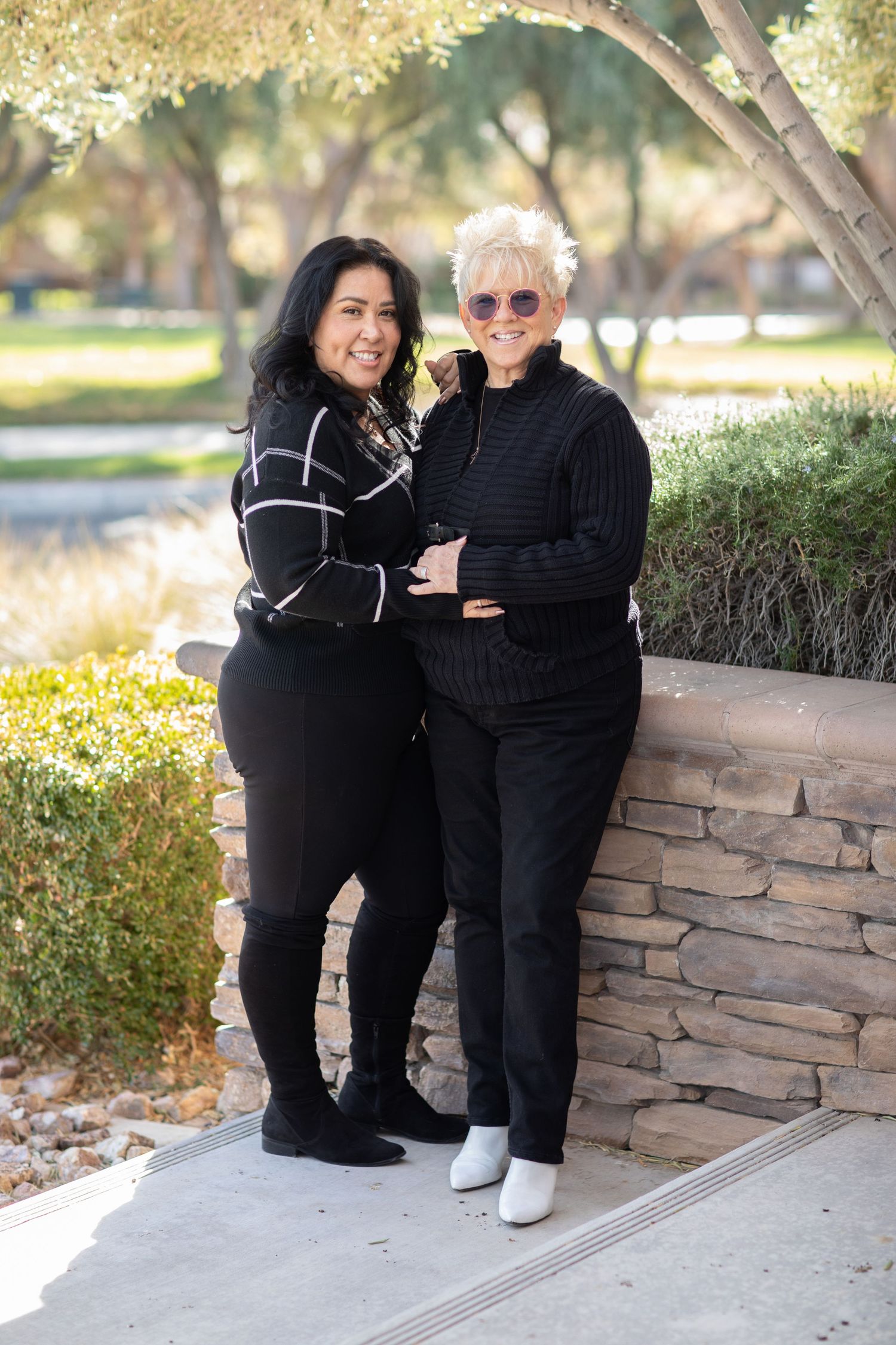 Two people in black outfits smiling together in sunny park setting.