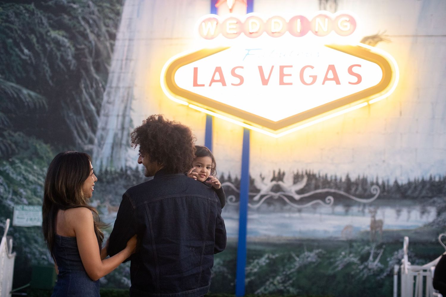 Two people stand in front of an illuminated Las Vegas welcome sign against a moody nature backdrop.