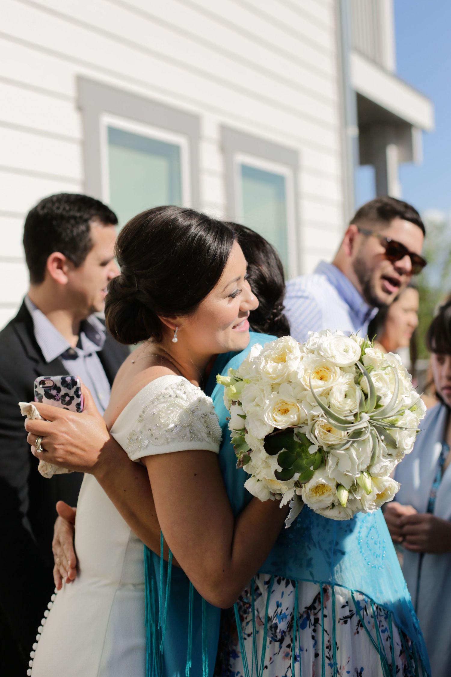 A person in a wedding dress holds a beautiful white bouquet while standing with guests outdoors.