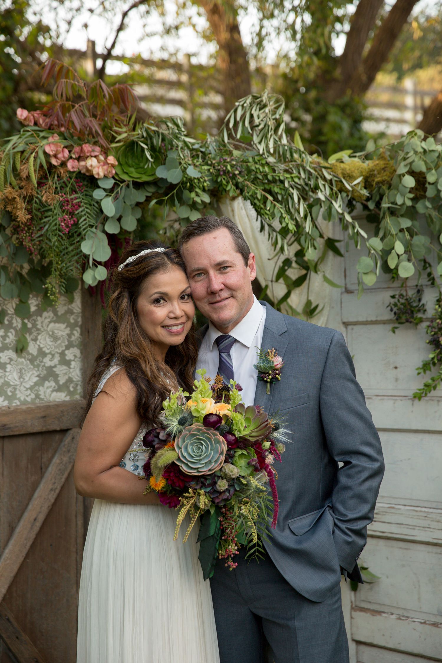 A wedding couple poses in front of a rustic wooden wall with greenery and floral decor.