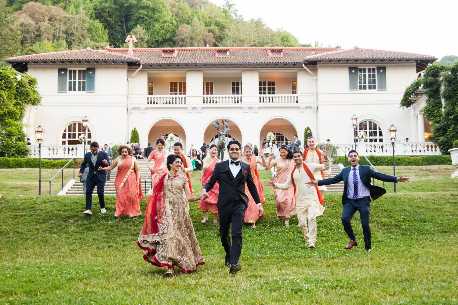 Wedding party jumps for joy outside a white mansion during celebration photo shoot.