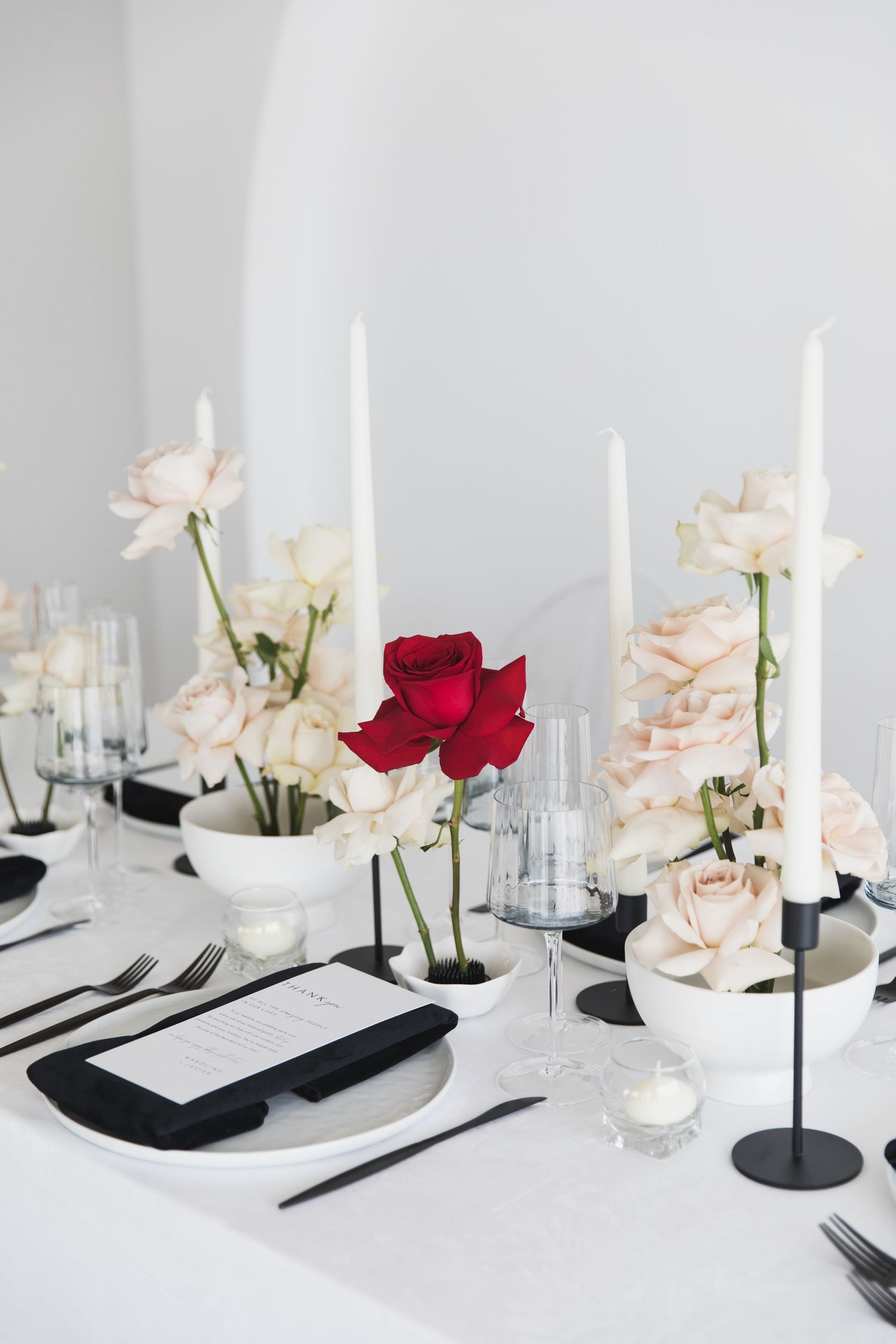 Elegant table setting with white candles, blush roses, and a single red rose centerpiece on crisp white linens.