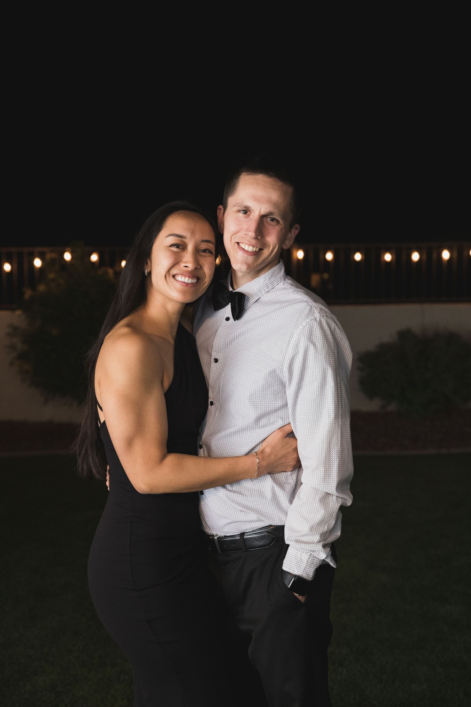 Formal couple embraces at night under string lights in black dress and white shirt.