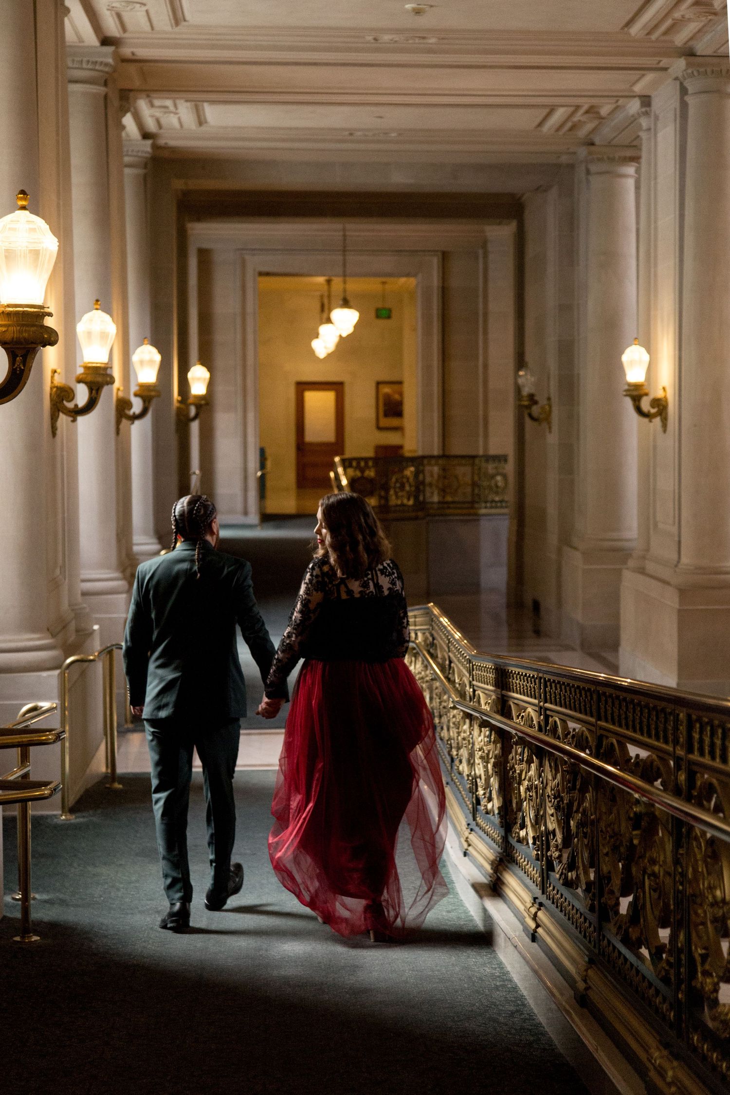 A couple walking down an elegant hallway with ornate gold railings and warm lighting.
