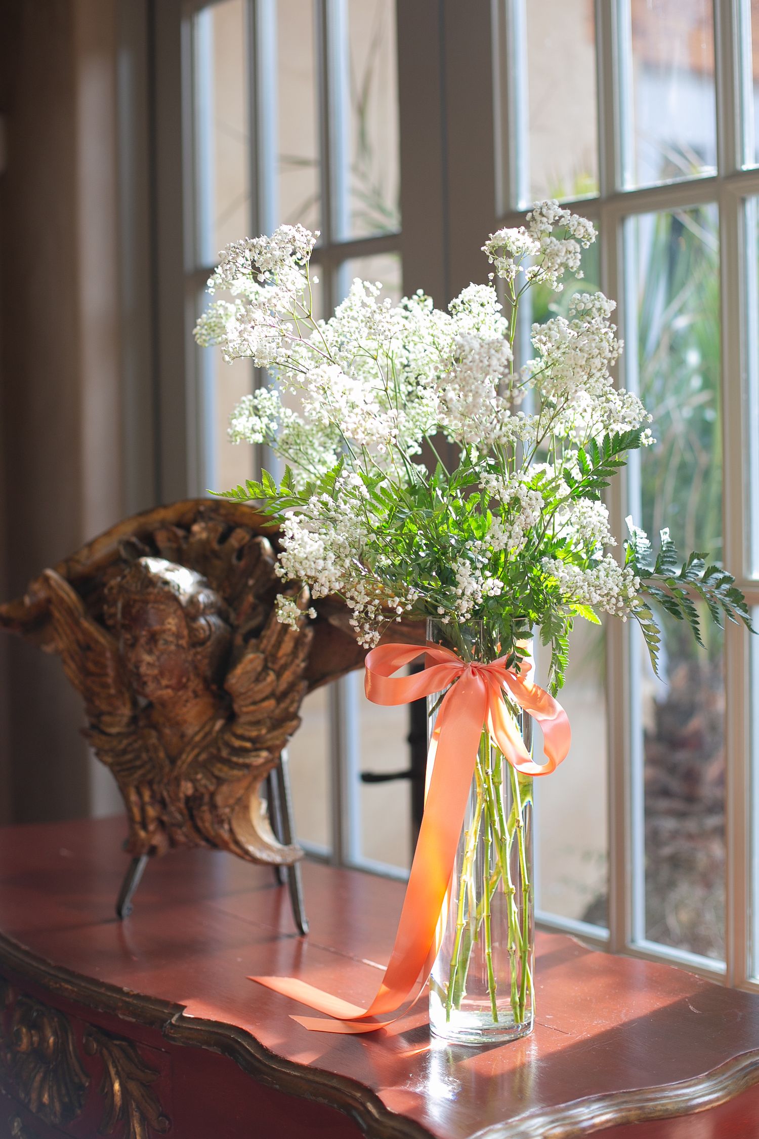 Delicate white baby's breath flowers in a glass vase with coral ribbon near a window.