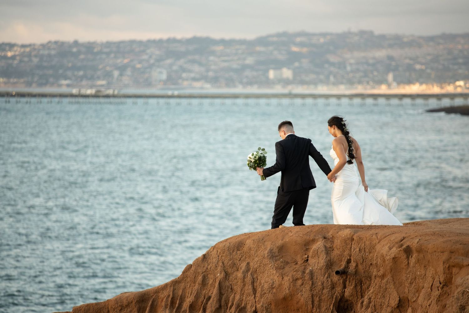 A couple stands on coastal rocks overlooking the ocean in wedding attire.